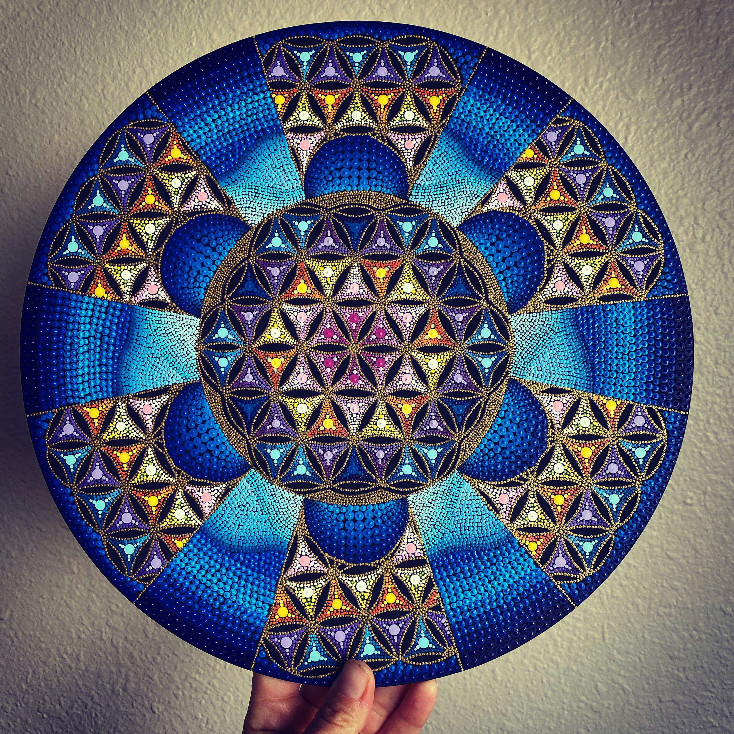 Colorful painted circular artwork featuring intricate geometric patterns with blue, purple, yellow, pink, and white dots, held by a person's hand against a beige wall.