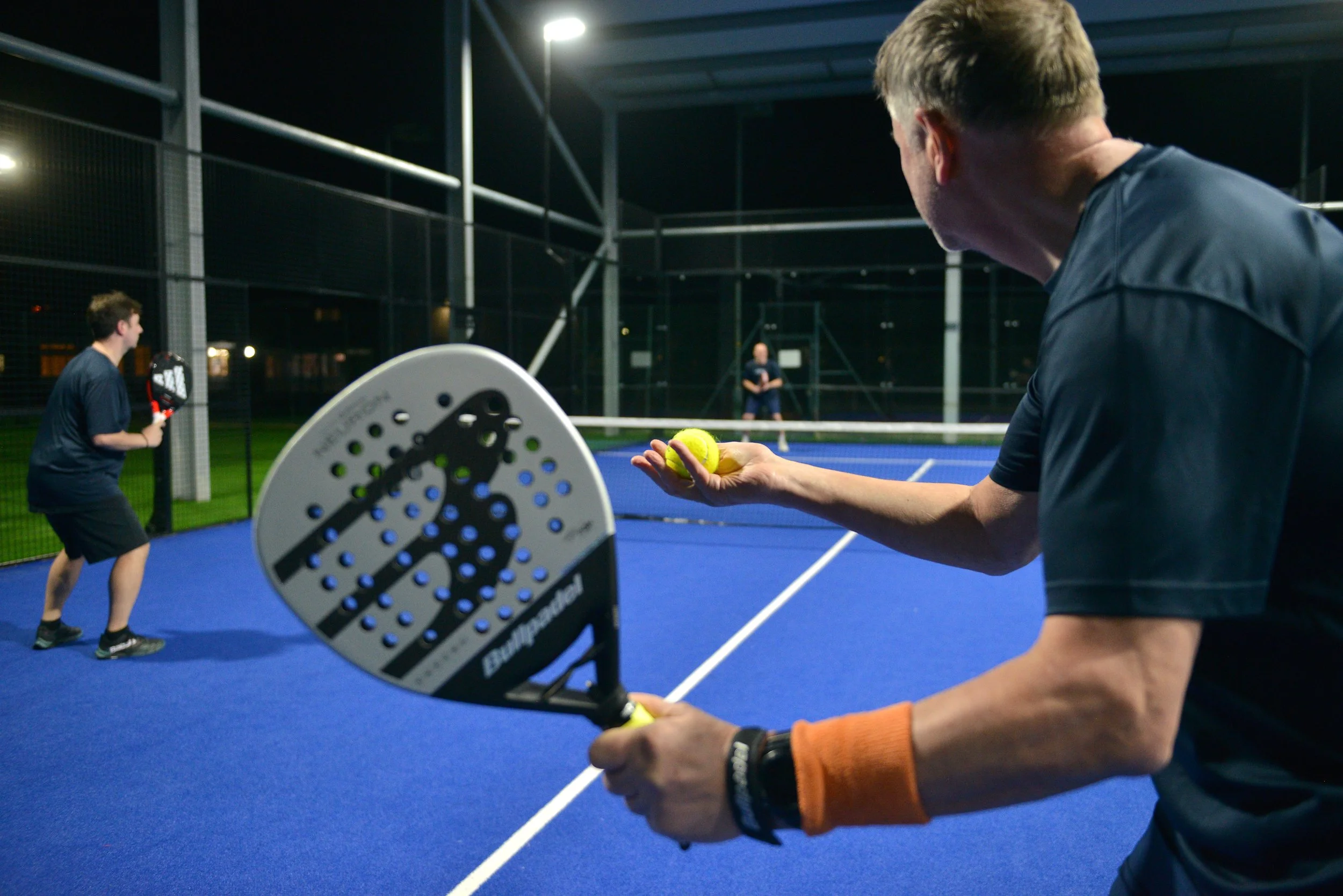 Two men playing pickleball on an outdoor court at night, one is holding a paddle and the other is holding a tennis ball, with a fence and lighting in the background.
