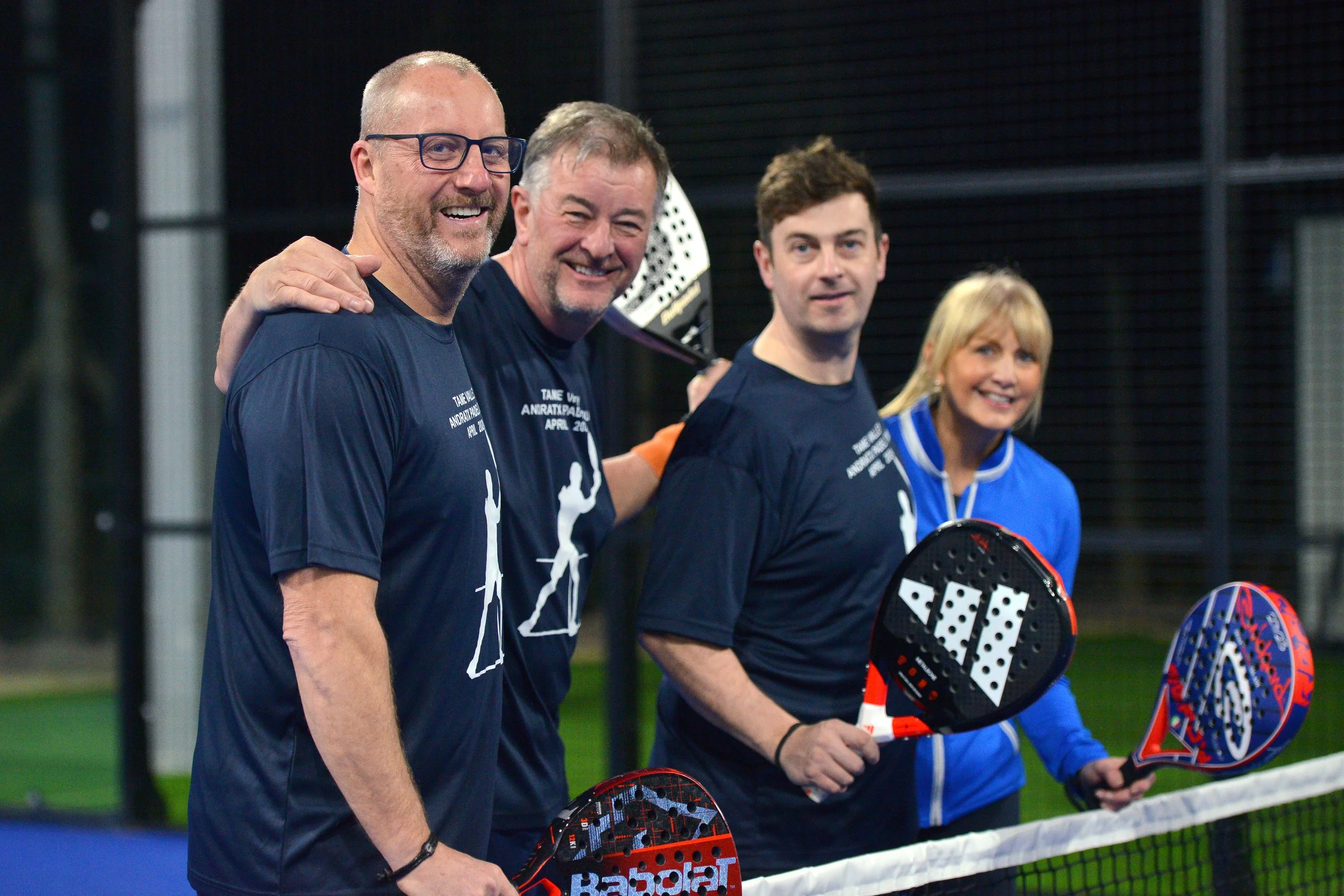 Four people, three men and one woman, stand on a pickleball court holding paddles, smiling. They are dressed in sports attire, with the men wearing matching T-shirts, and the woman in a blue jacket.