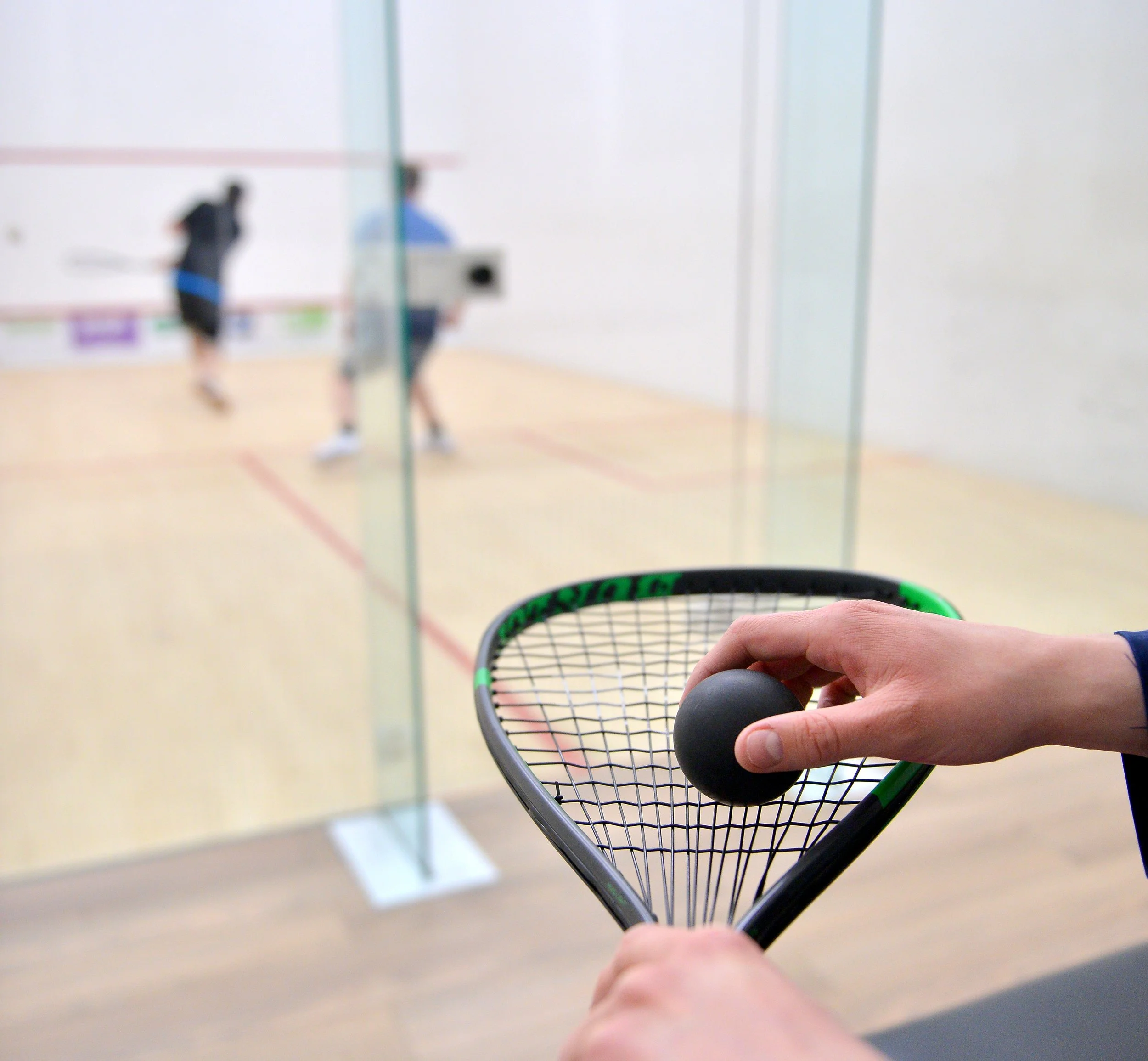 A person holding a squash racket with a black ball in front of a squash court where two players are playing in the background.