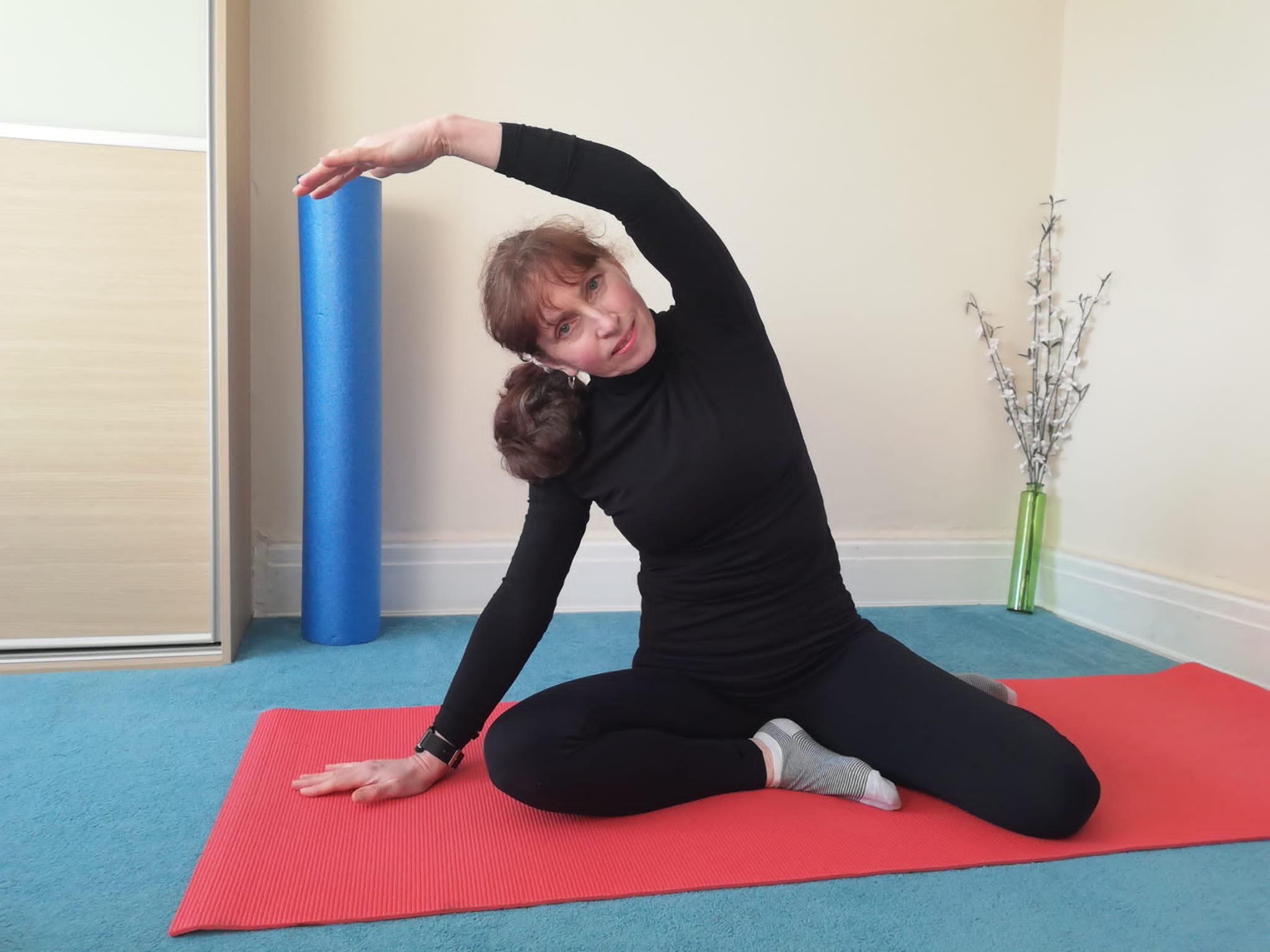 A woman practicing yoga on a red mat indoors, performing a side stretch with one arm extended overhead and the other hand on the mat, wearing a black long-sleeved shirt and black pants.