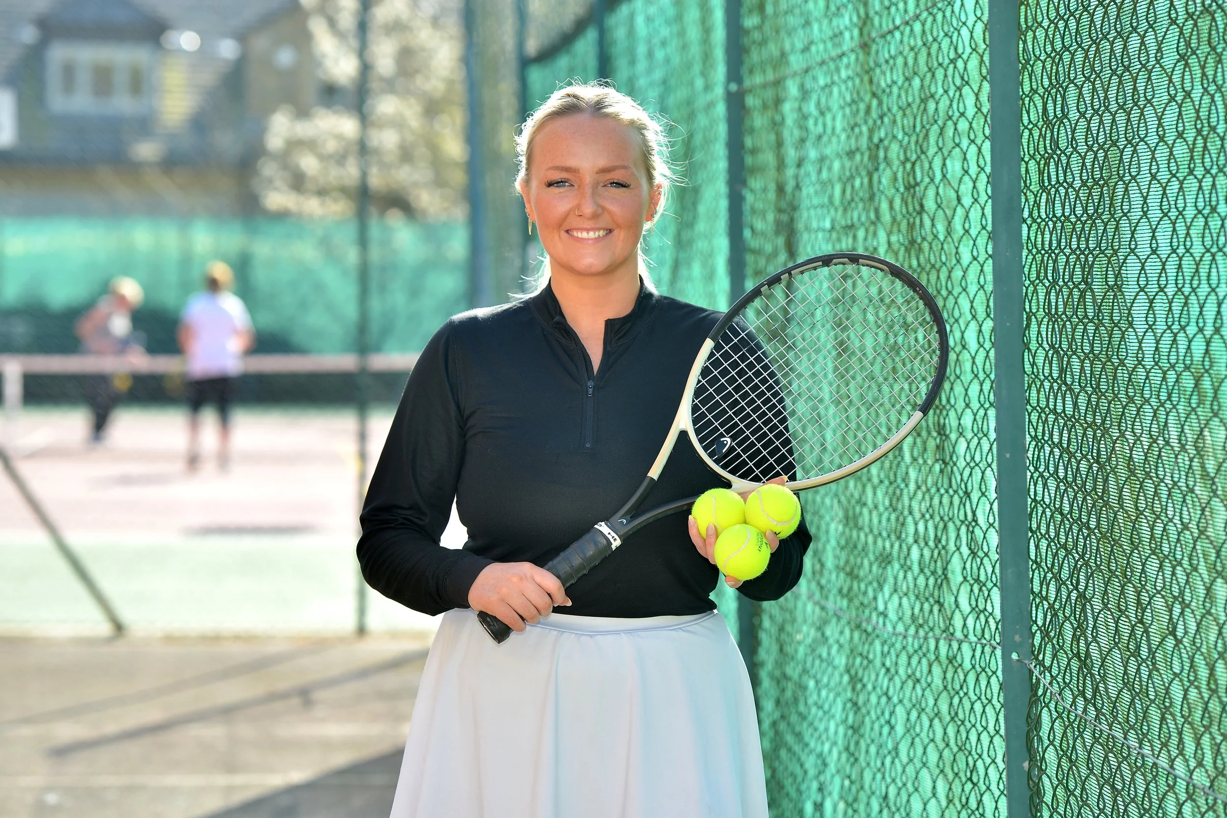 A woman standing on a tennis court holding a tennis racket and four tennis balls, smiling at the camera.