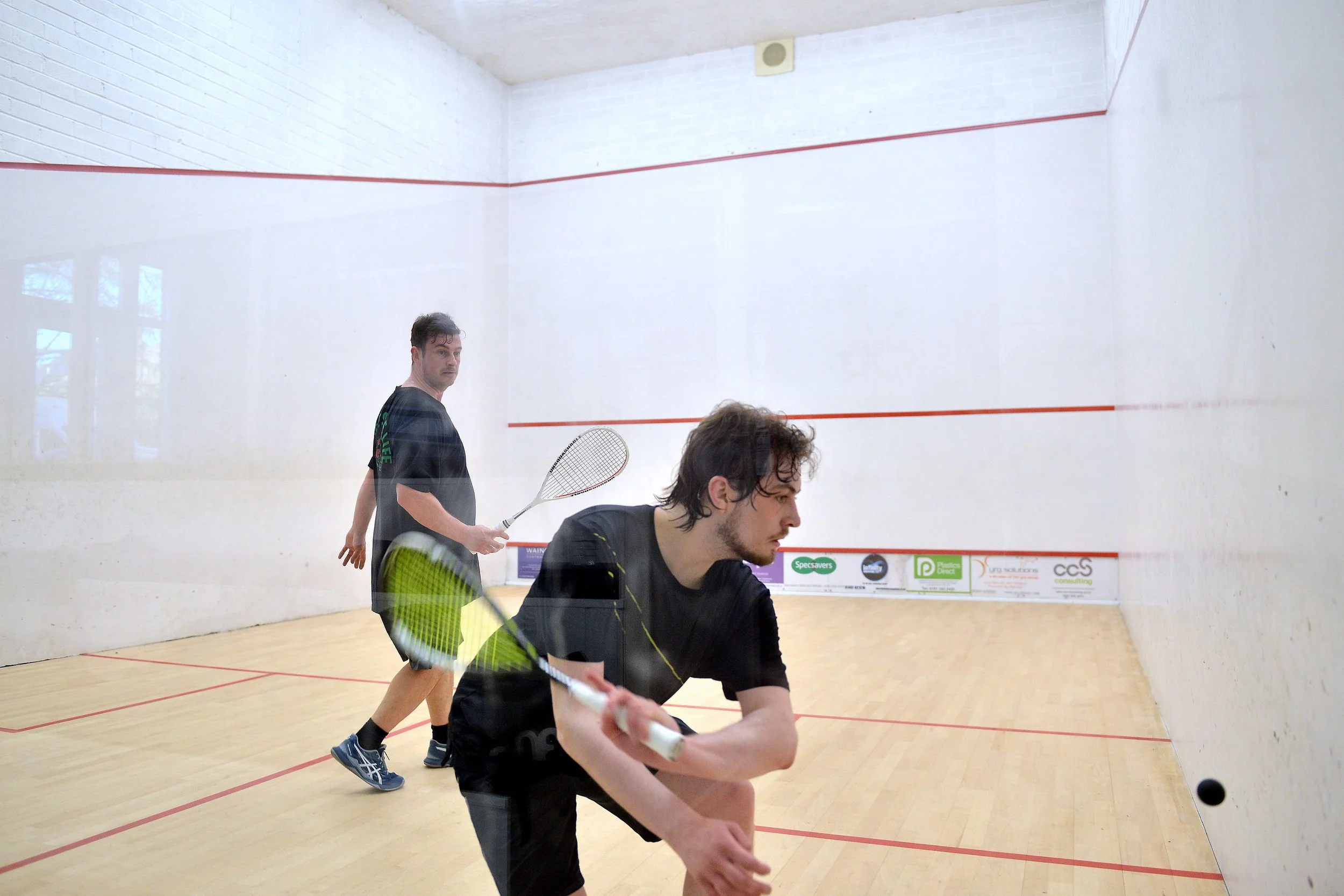 Two men playing squash on an indoor court with white walls and a wooden floor, one preparing to hit the ball and the other standing behind.