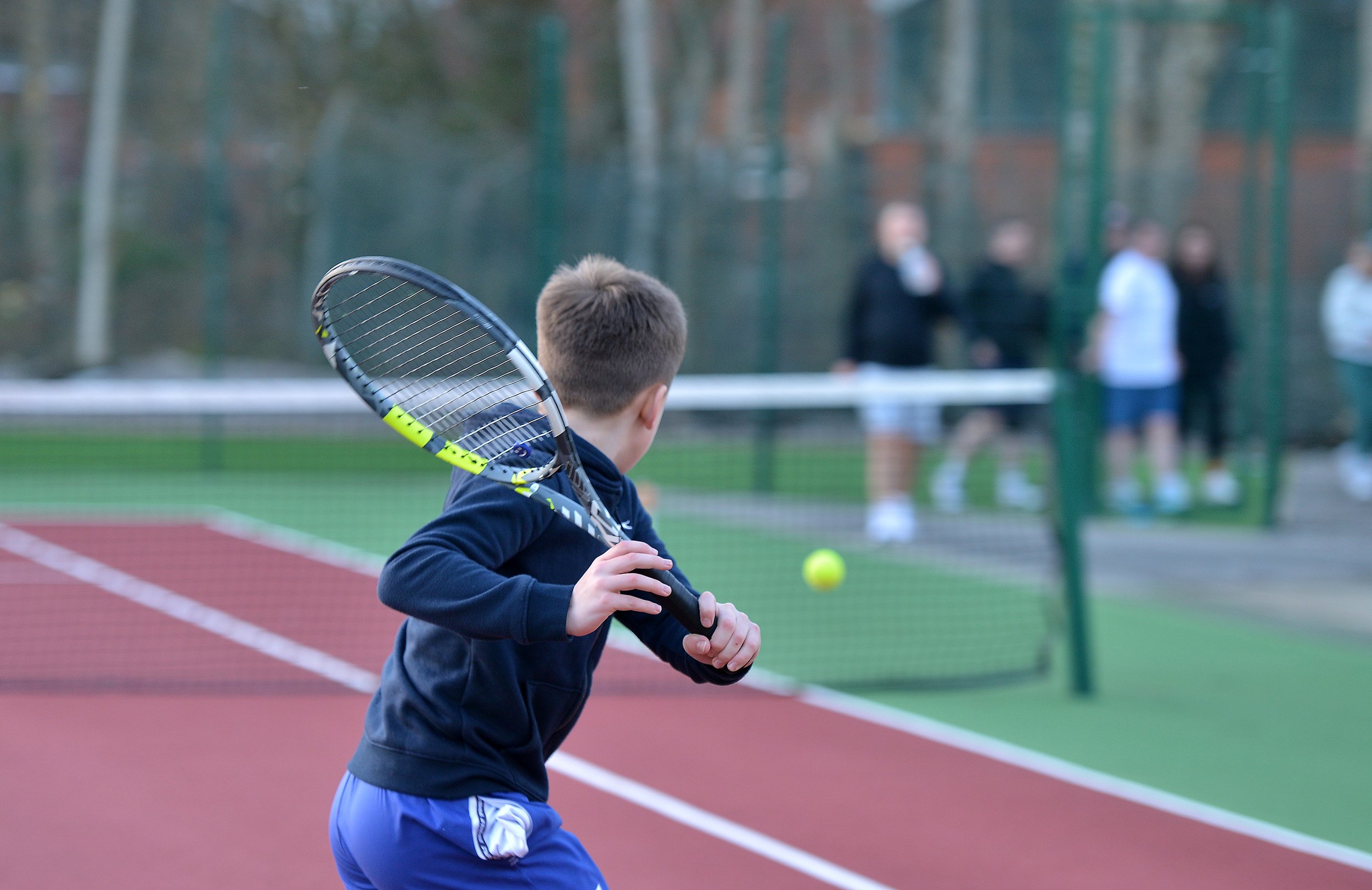A young boy playing tennis on an outdoor court, preparing to hit a tennis ball with a racket. In the background, several people are watching near the fence.