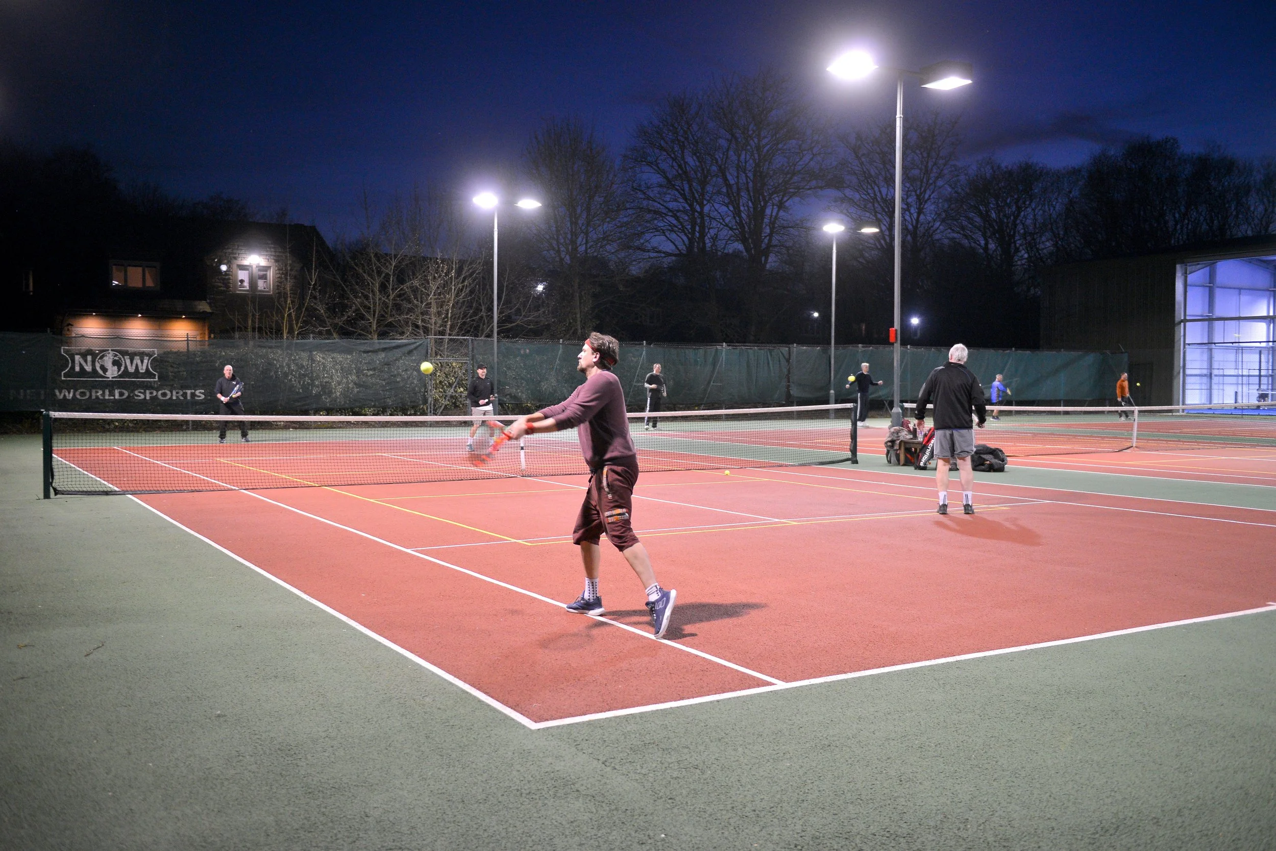People playing tennis on an outdoor court at night under bright lights, with trees and a building in the background.