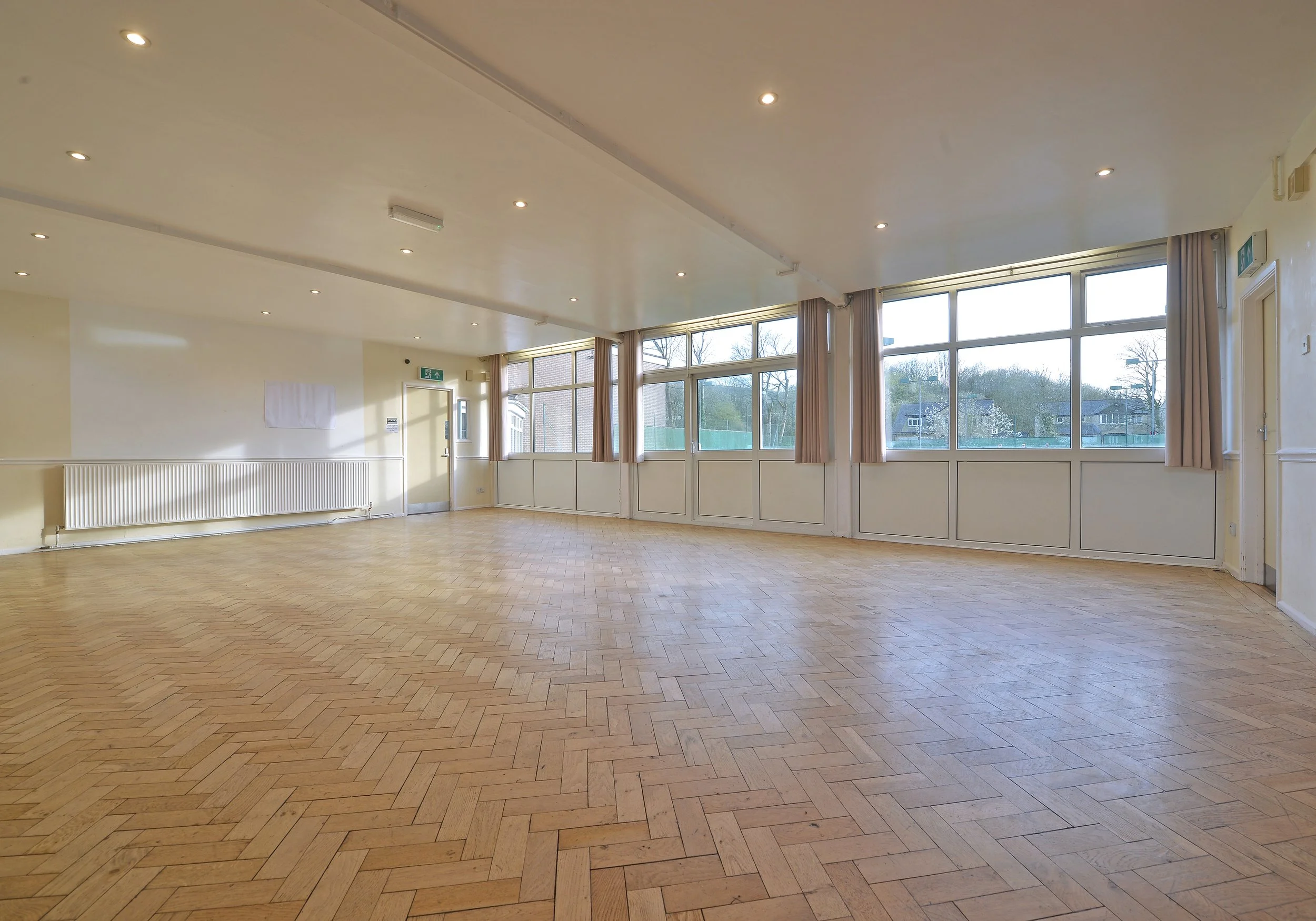 Empty room with large windows, wooden herringbone floor, beige curtains, and ceiling lights.