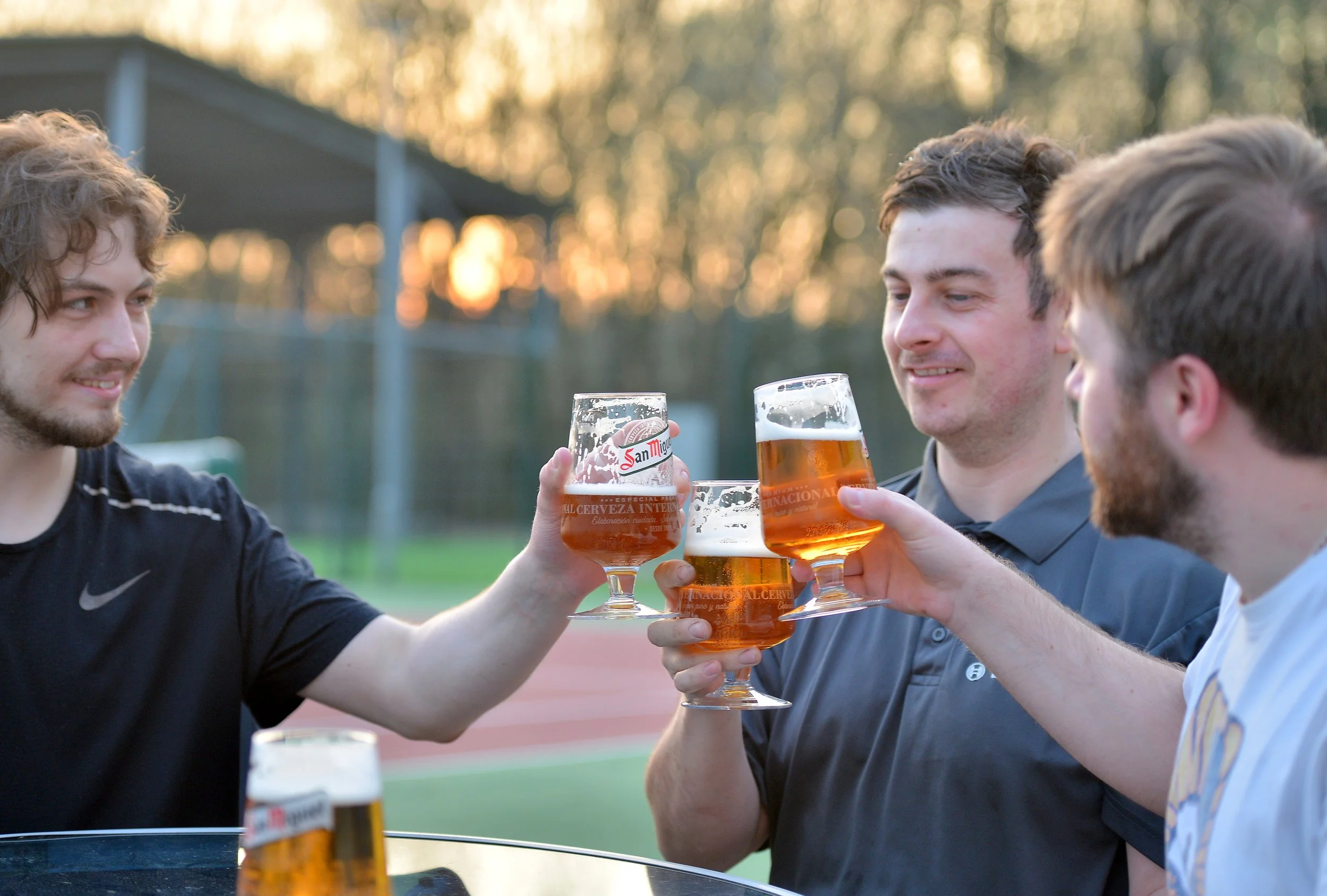 Three men clinking glasses of beer in an outdoor setting during sunset.