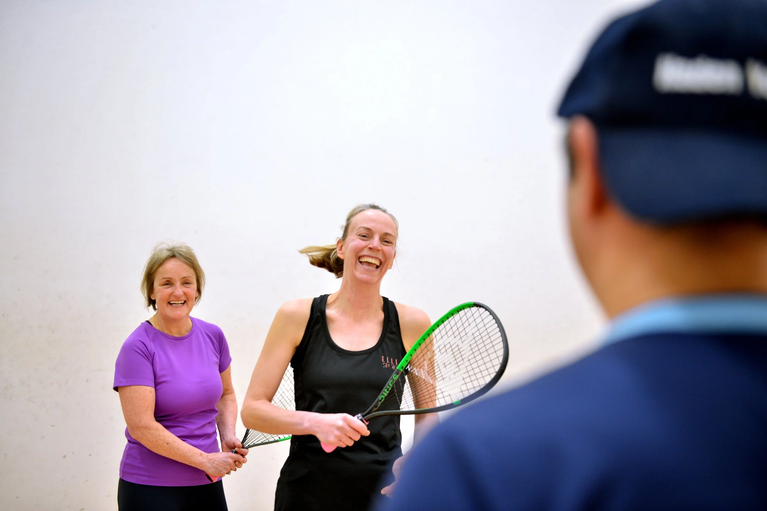 Two women and a man playing indoor racquetball, with the women smiling and holding racquets, the man wearing a blue cap and the women dressed in athletic clothing.