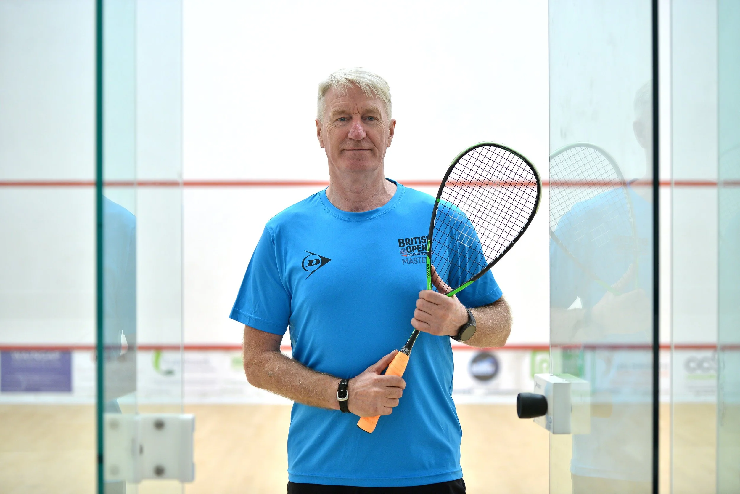 A man in a blue T-shirt holding a squash racket standing in front of the glass wall of an indoor squash court.