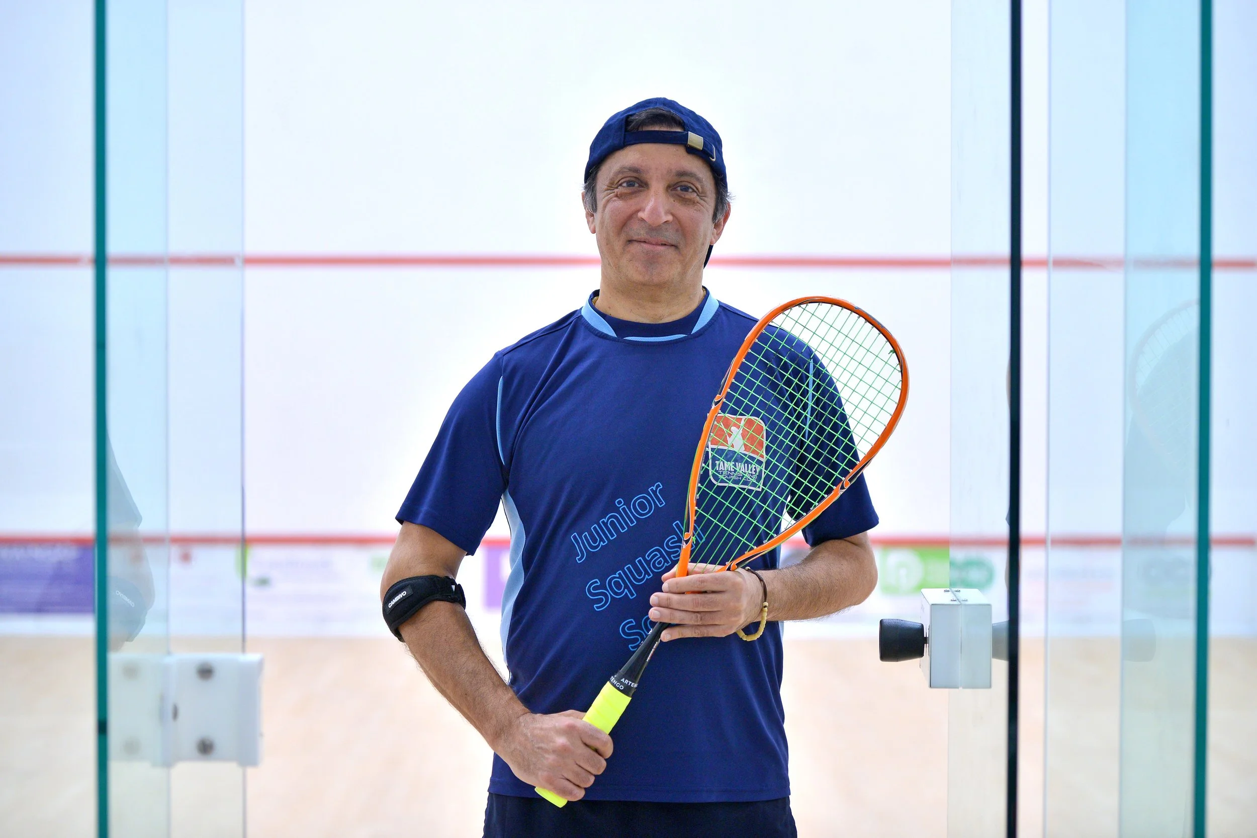 A man in a blue sports shirt and cap holding a squash racket standing in front of an indoor squash court.