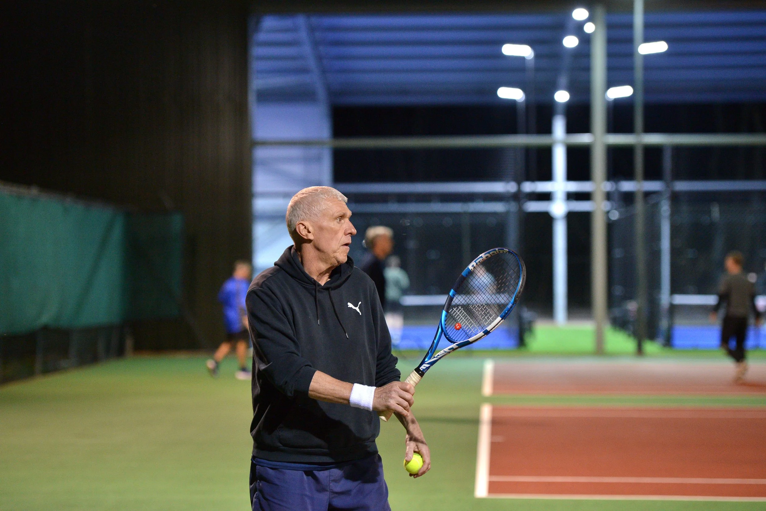 An elderly man standing on a tennis court, holding a tennis racket and a tennis ball, with several other players practicing in the background at night.