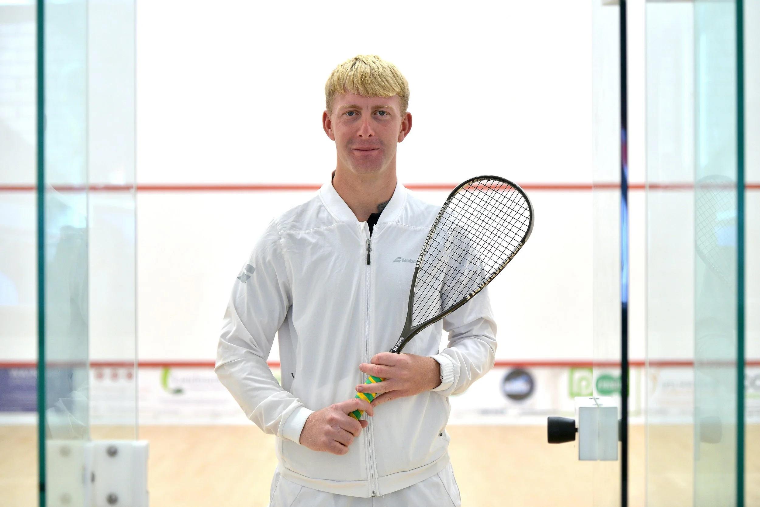 A young man in a white sports jacket holding a squash racket standing on an indoor squash court.