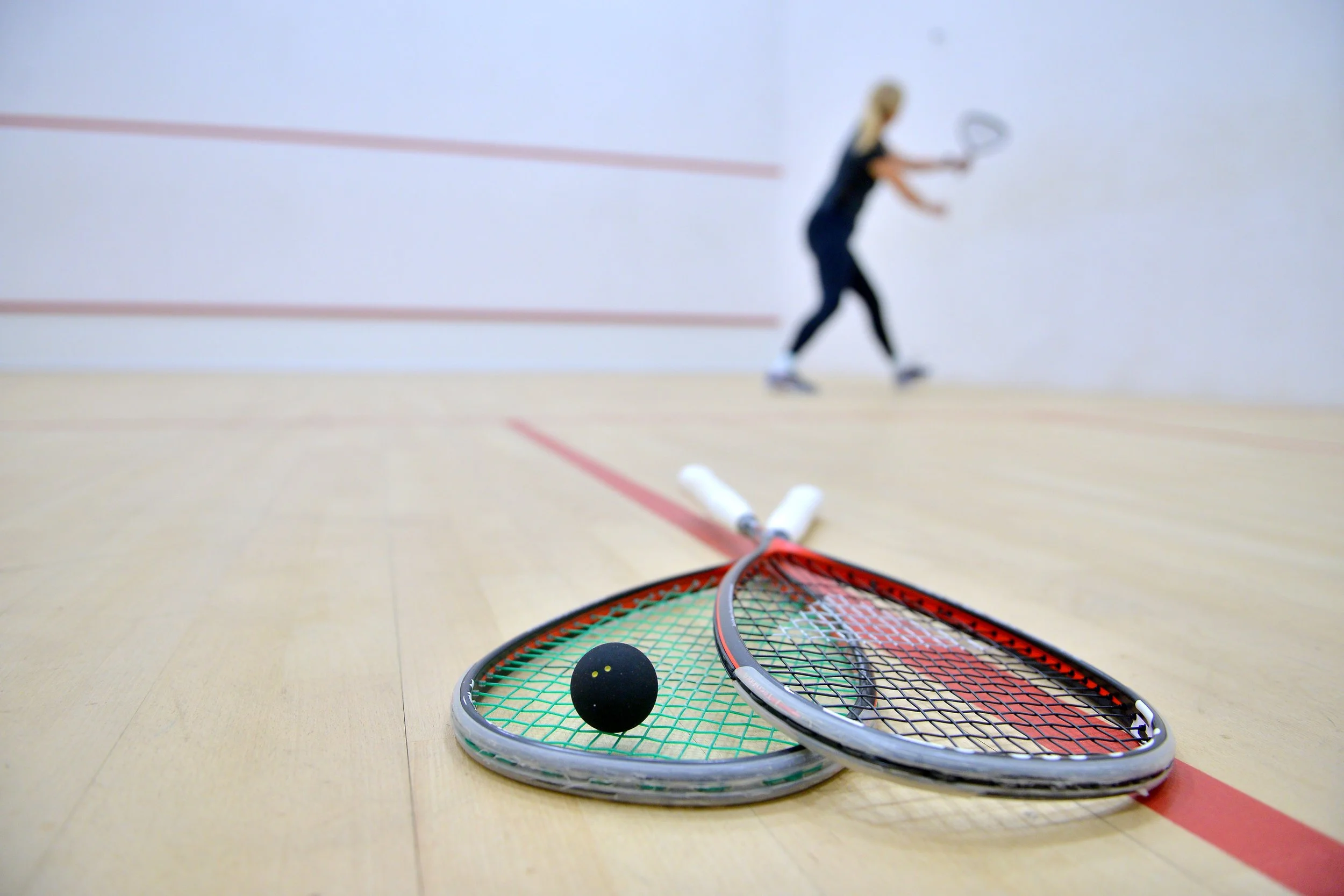 Two squash rackets and a black squash ball on the court with a woman holding a racket in the background.