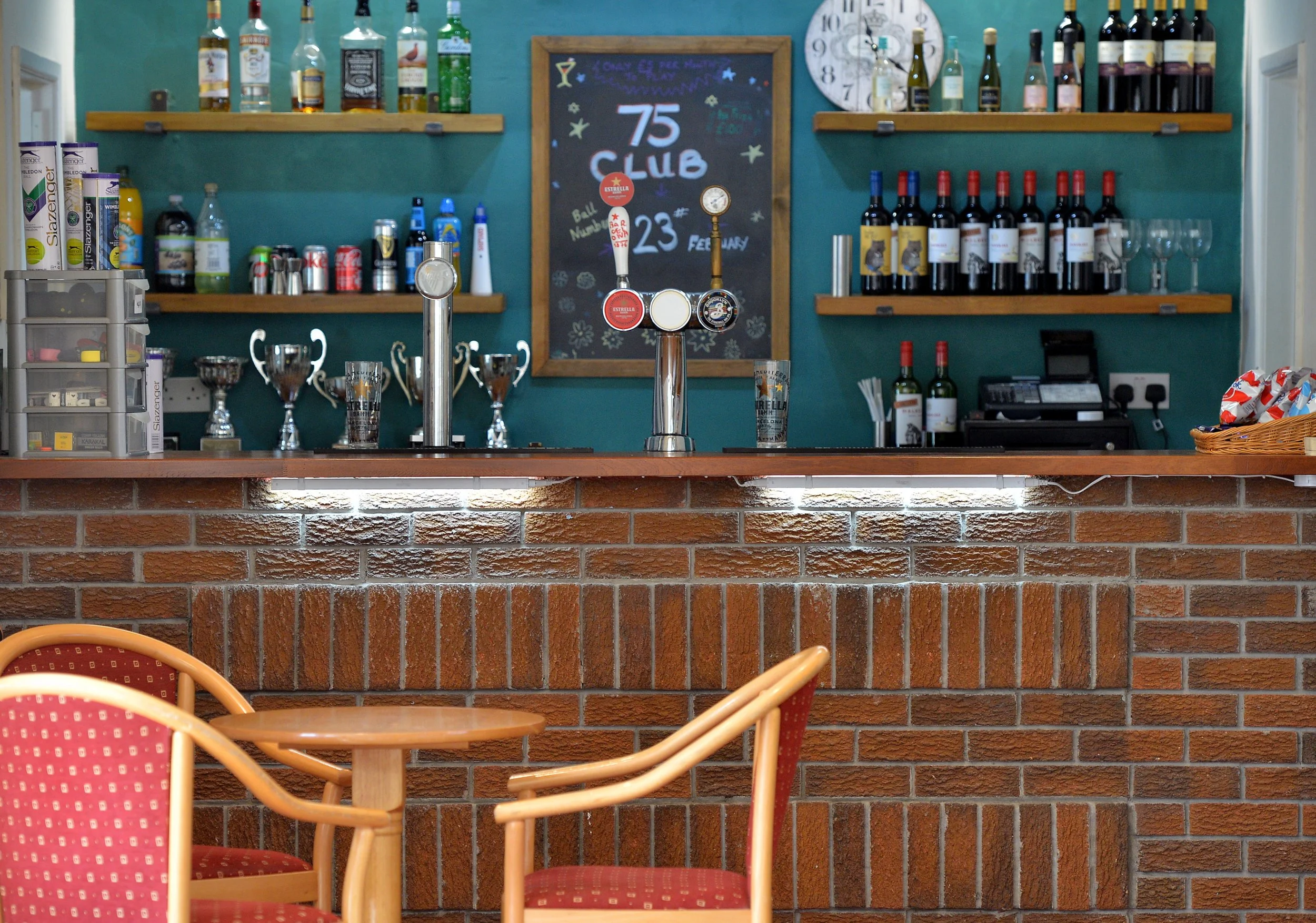 A bar with a brick front, wooden countertop, and colorful chairs. Behind the bar, there are shelves with various bottles of alcohol, trophies, a chalkboard with handwritten notes, and a clock on the wall.