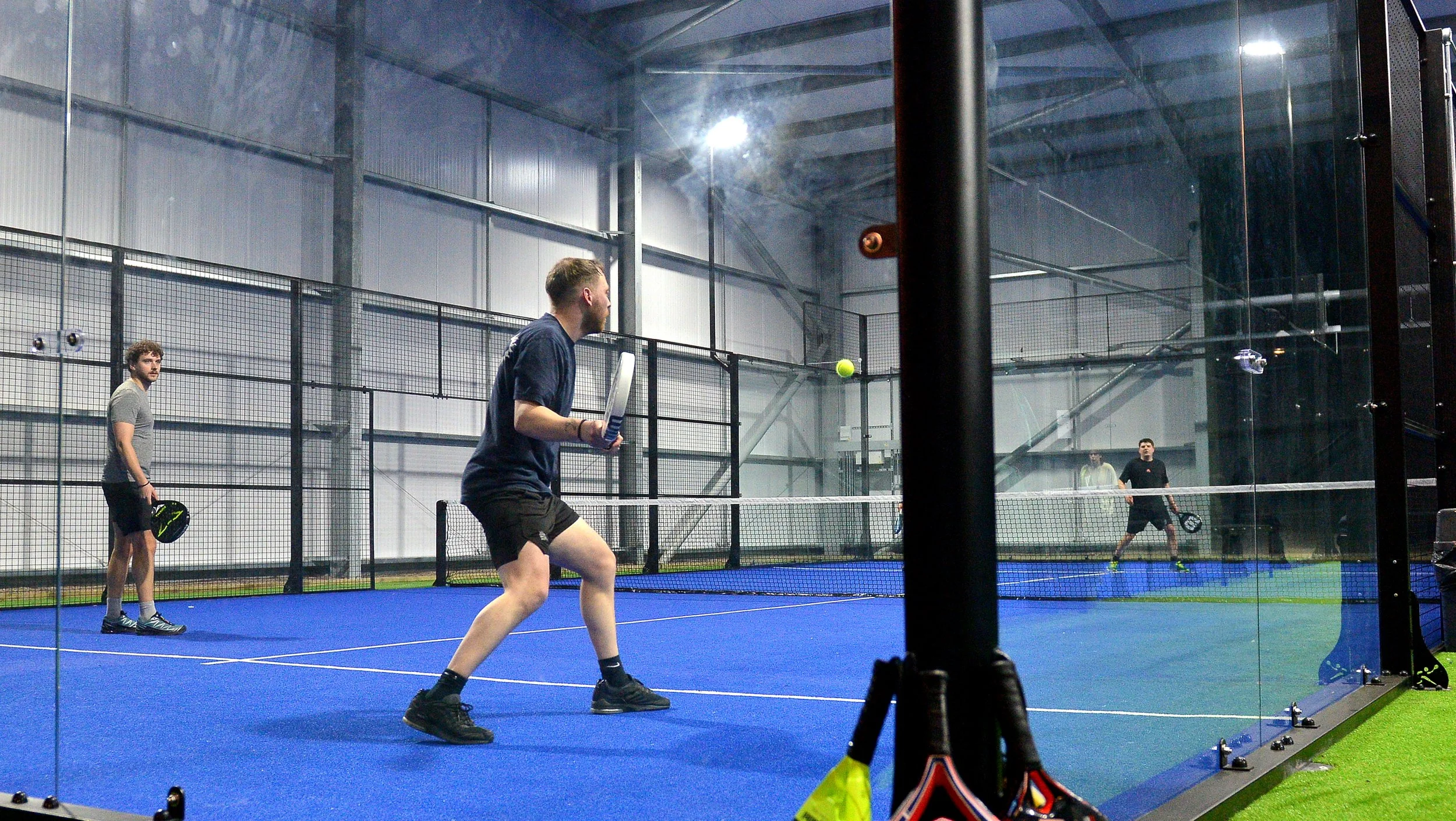 Four men playing pickleball on an indoor court with blue flooring, with a net in the middle and a glass wall on the right side.