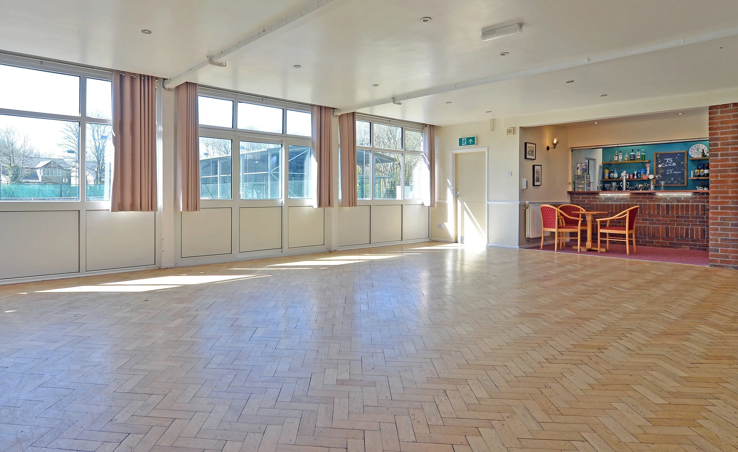 Empty spacious room with large windows, wooden flooring, and a small bar area with red chairs and a brick counter.