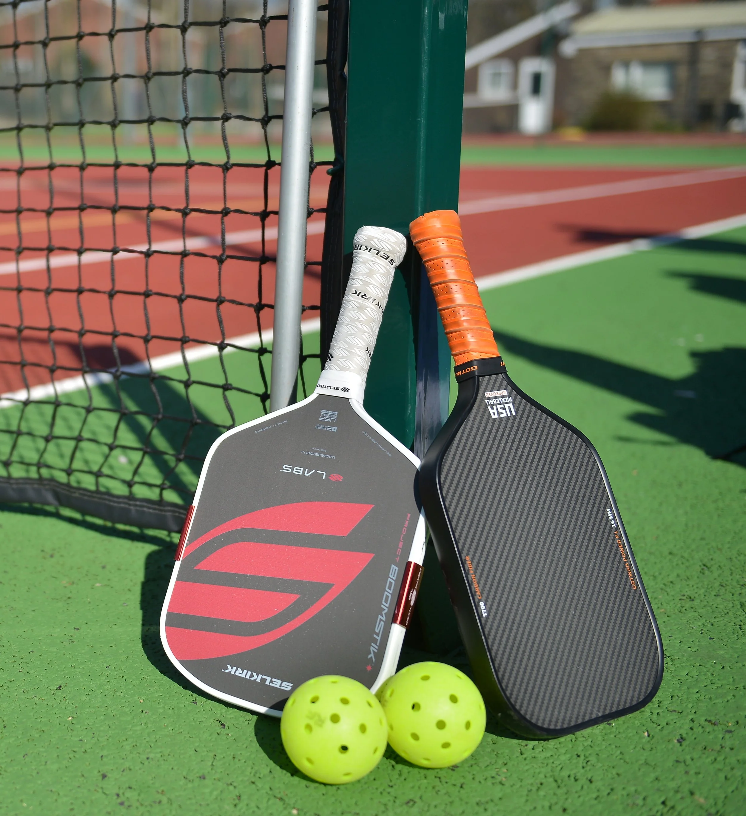Two pickleball paddles leaning against a green pole near a tennis court, with two yellow pickleballs in front.