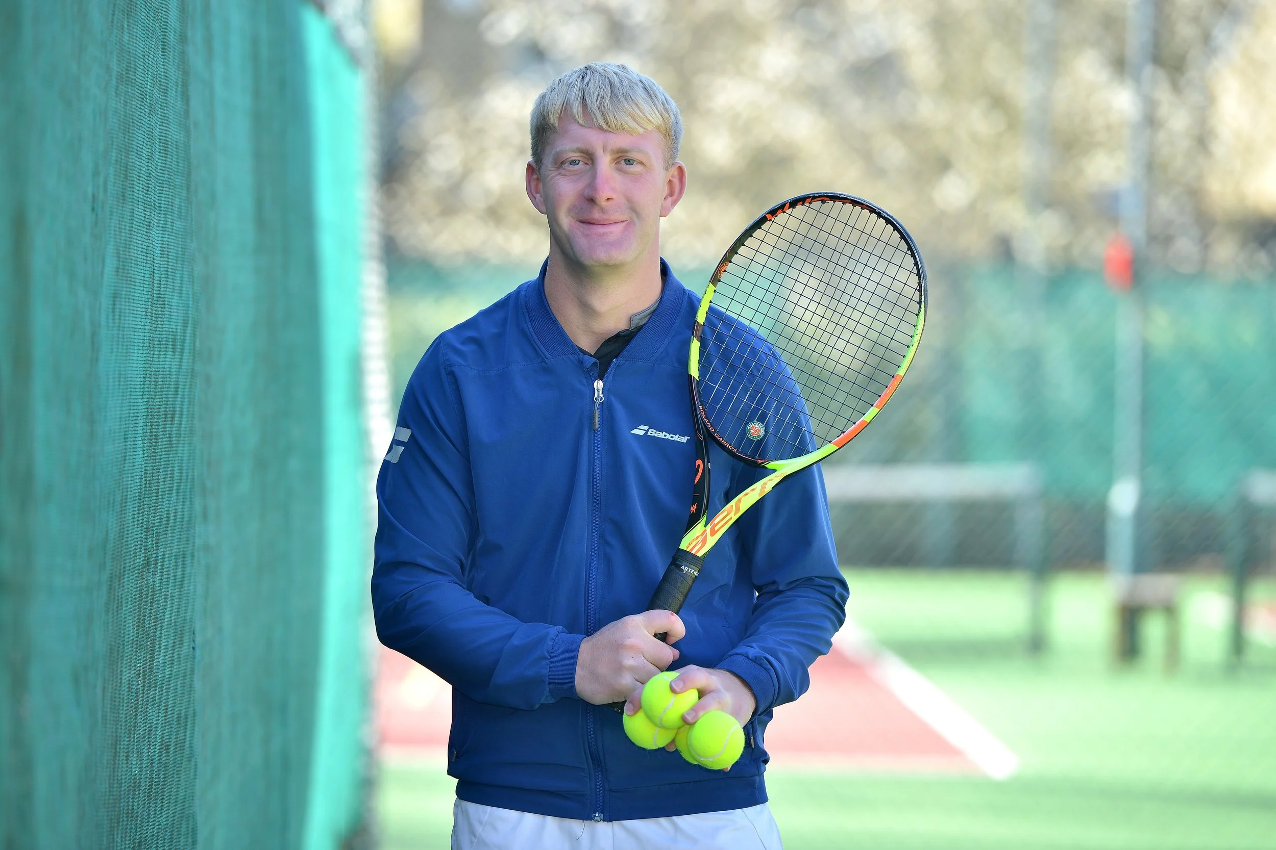 A young man in a blue sports jacket holding tennis rackets and tennis balls on outdoor tennis court.