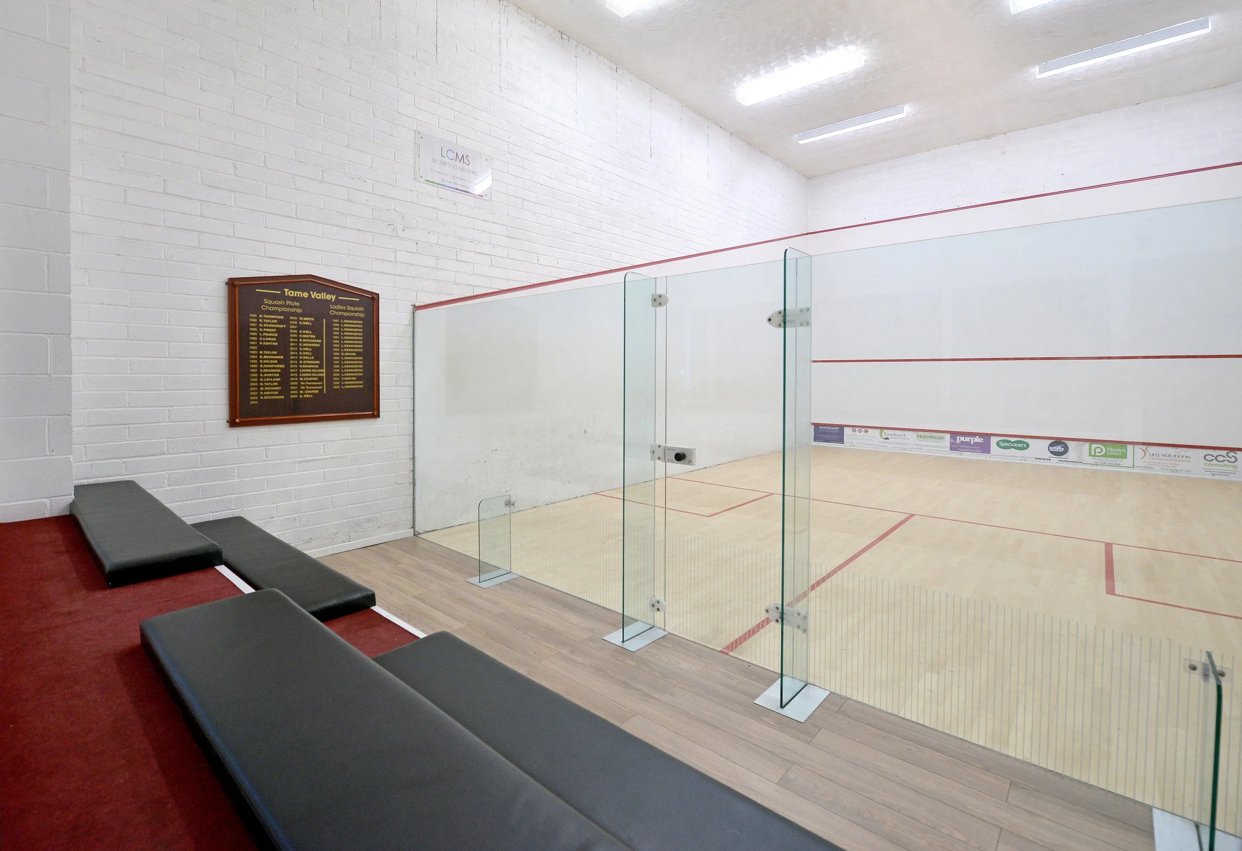 Indoor squash court with wooden floor, white brick wall, glass court enclosure, and black benches