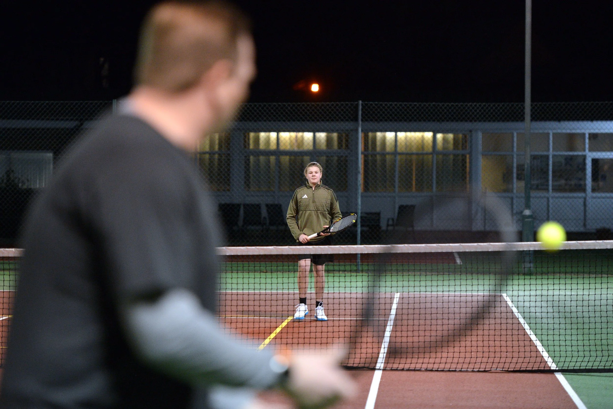 A girl holding a tennis racket on a tennis court at night, with a blurred man in the foreground and a tennis ball in motion.