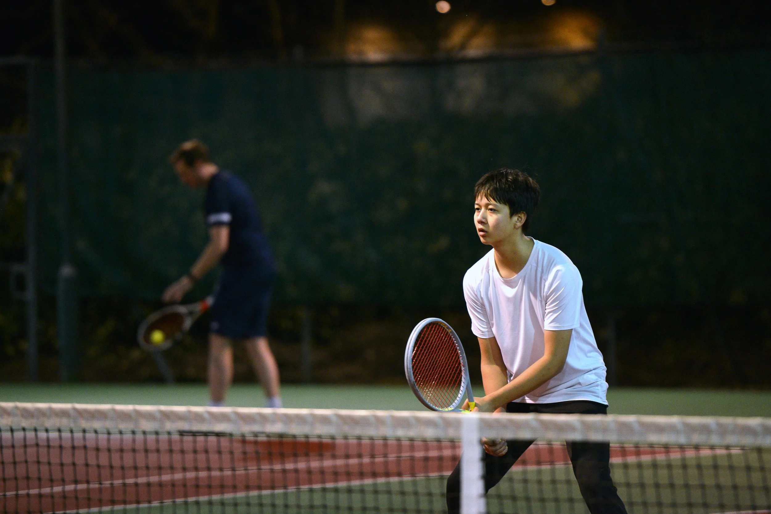 A young man in a white t-shirt holding a tennis racket on a tennis court, with another player in the background hitting a tennis ball.