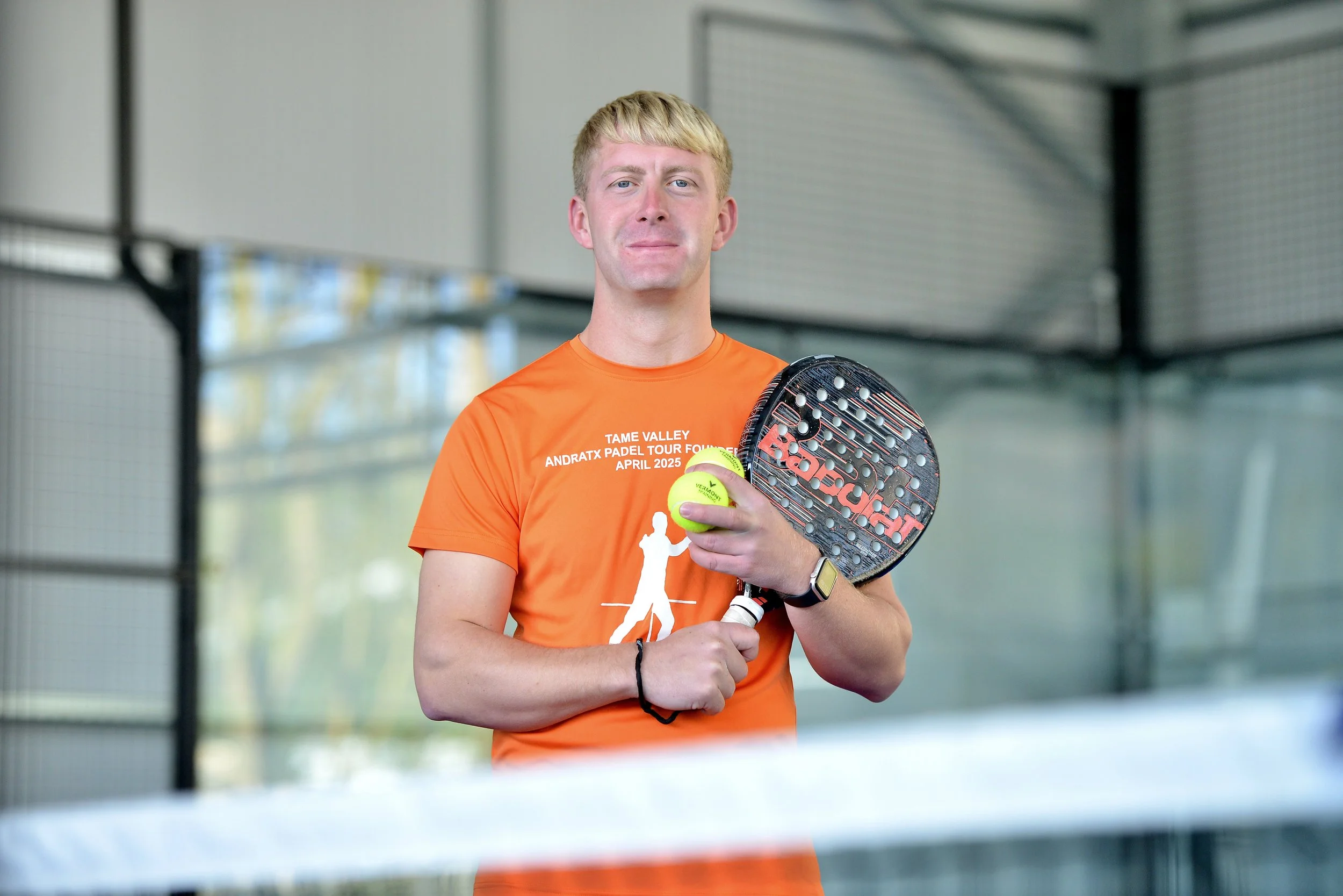 A man in an orange t-shirt holding a padel racket and a yellow padel ball inside an indoor padel court.