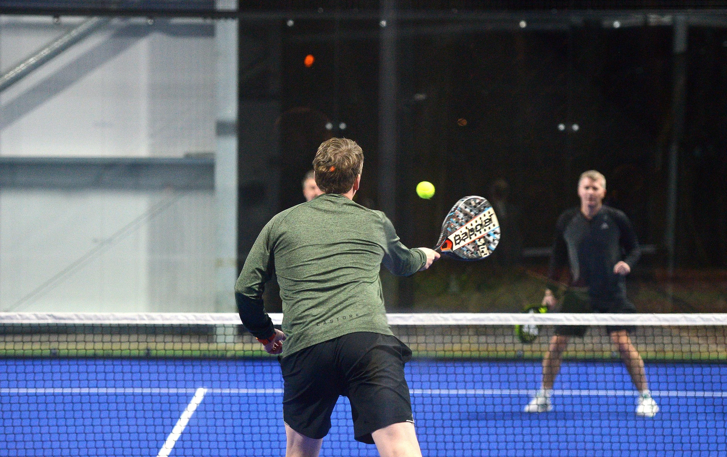 Two men playing pickleball on an outdoor court at night. The man in the foreground is about to hit a tennis ball with a paddle, while the other man is standing behind the net preparing to respond.
