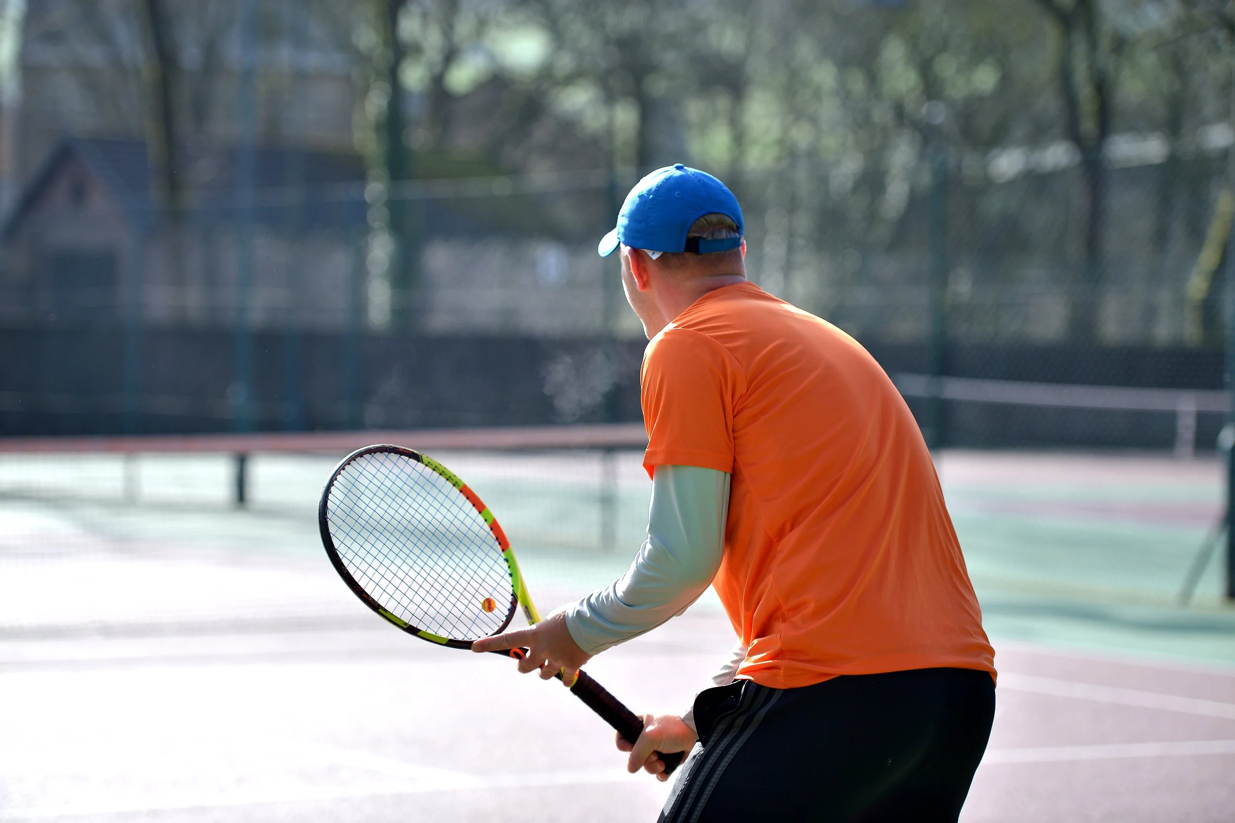 A person wearing an orange shirt, a blue cap, and black athletic shorts on an outdoor tennis court, holding a tennis racket.