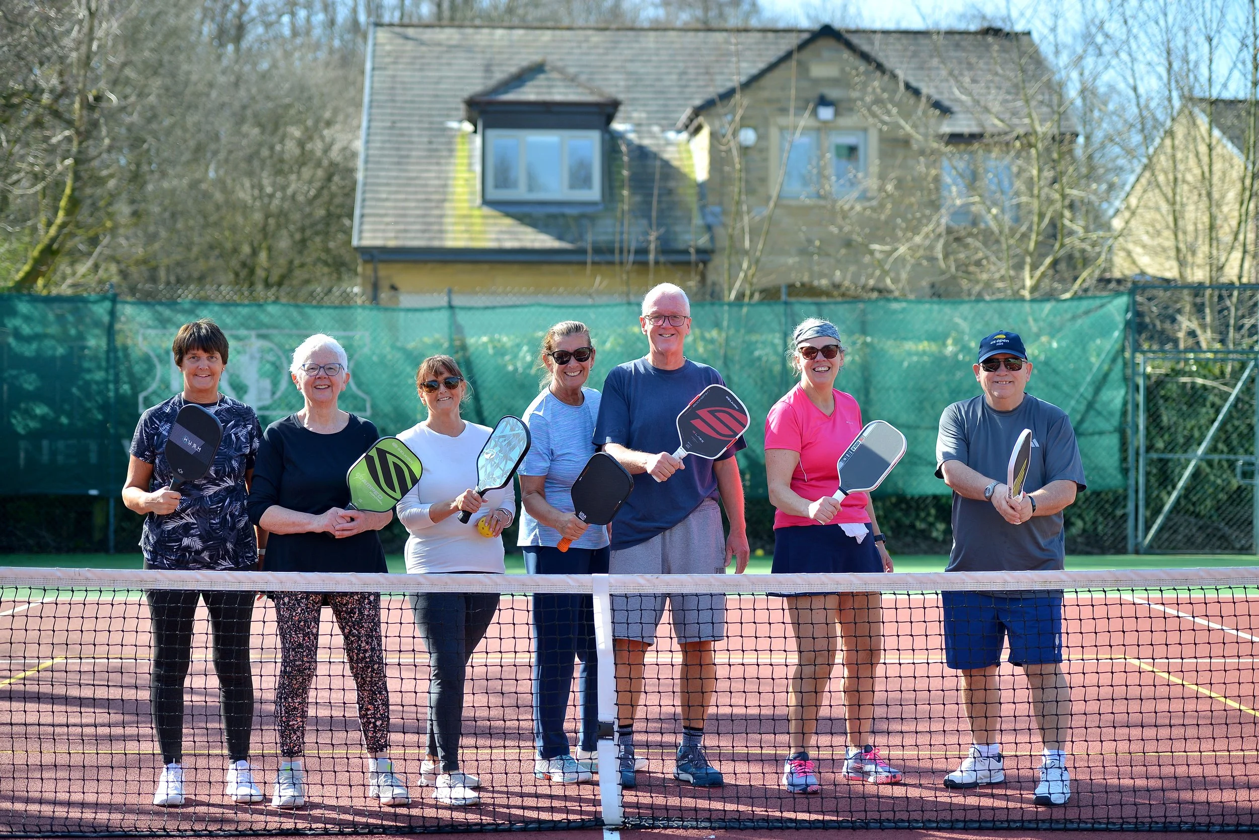 Group of seven people on a tennis court holding paddles, standing behind a net, with houses and trees in the background.