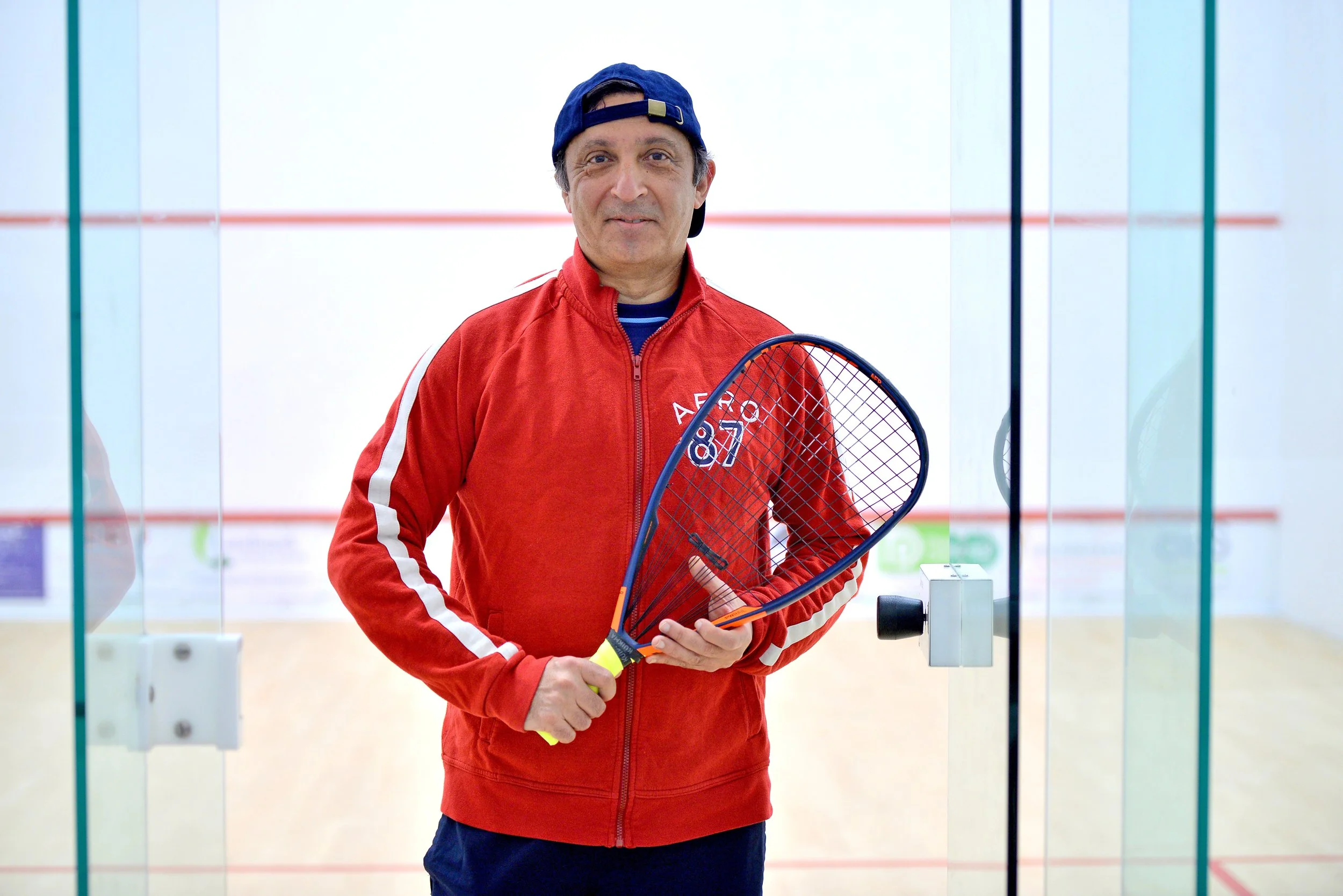 Man in red athletic jacket holding squash racket standing in squash court.