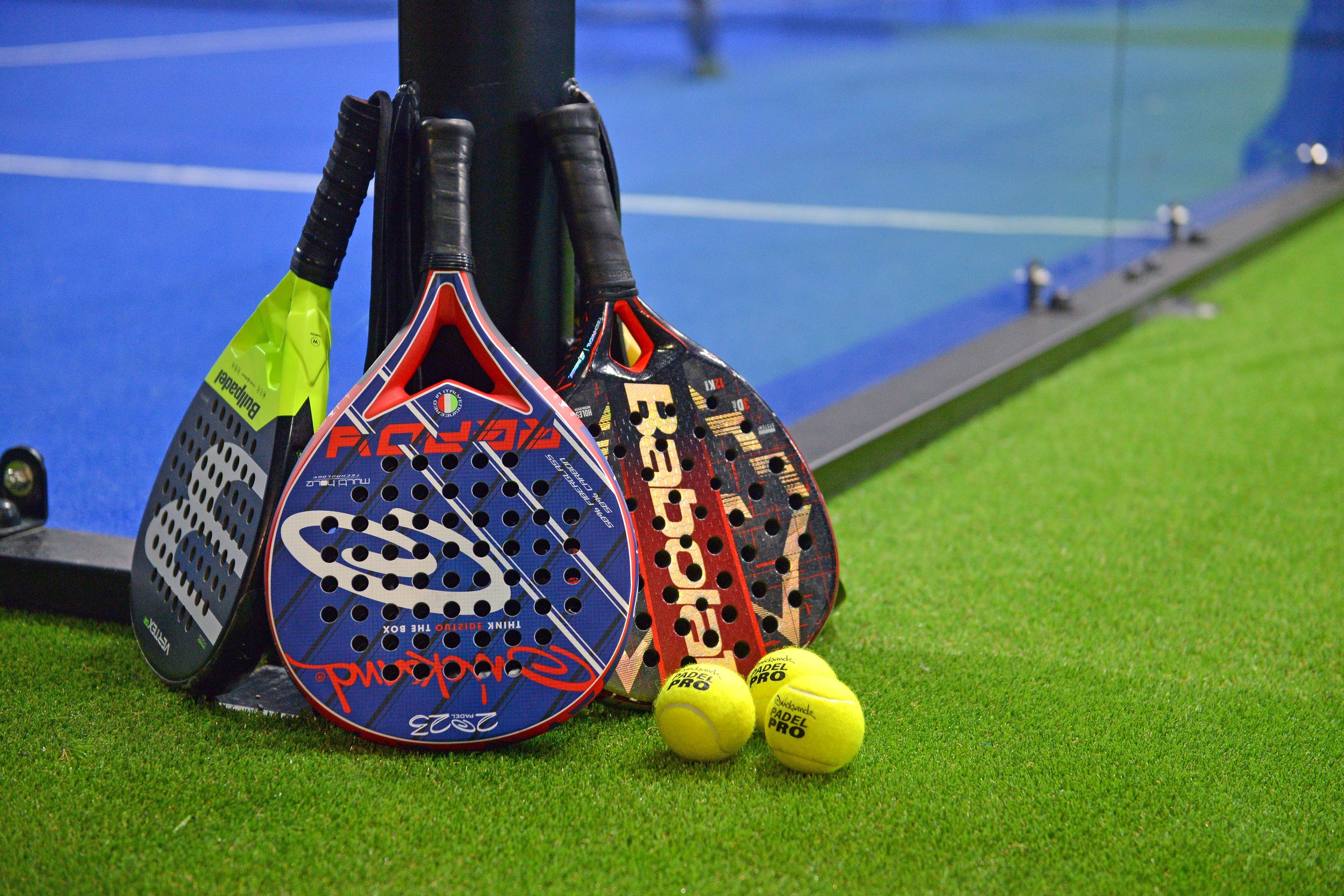 Padel rackets, yellow padel balls, and a green towel leaning against a black post on a green turf court.