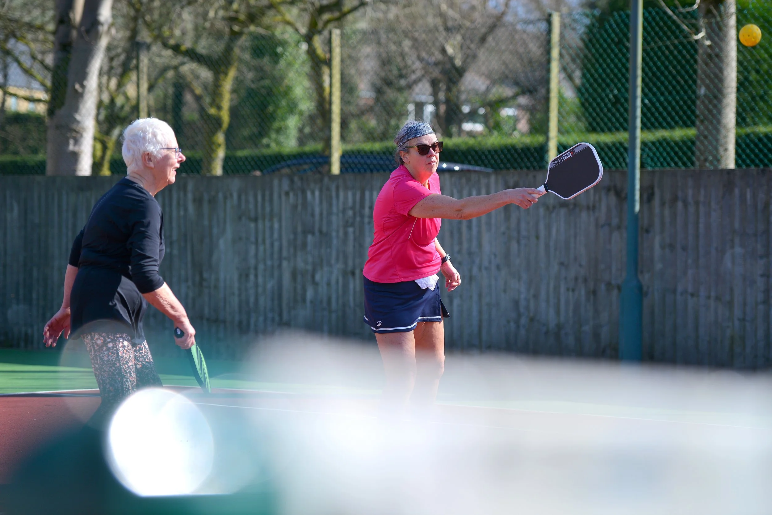 Two women playing tennis on an outdoor court during daytime. One woman in a pink shirt and shorts is preparing to hit the ball with a paddle, while the other woman in a black top and patterned pants watches.