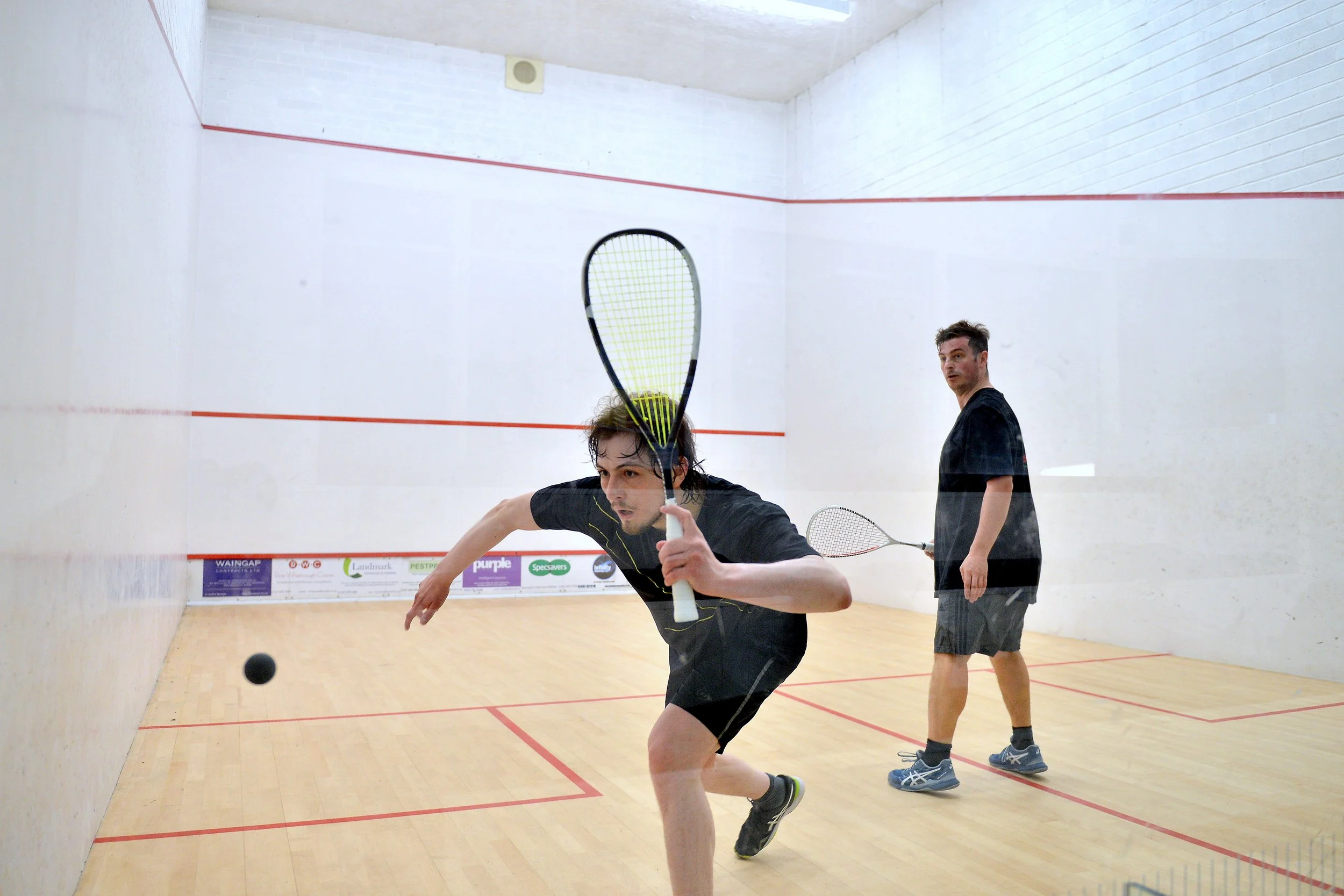 Two men playing squash inside a court, seen through a circular opening.