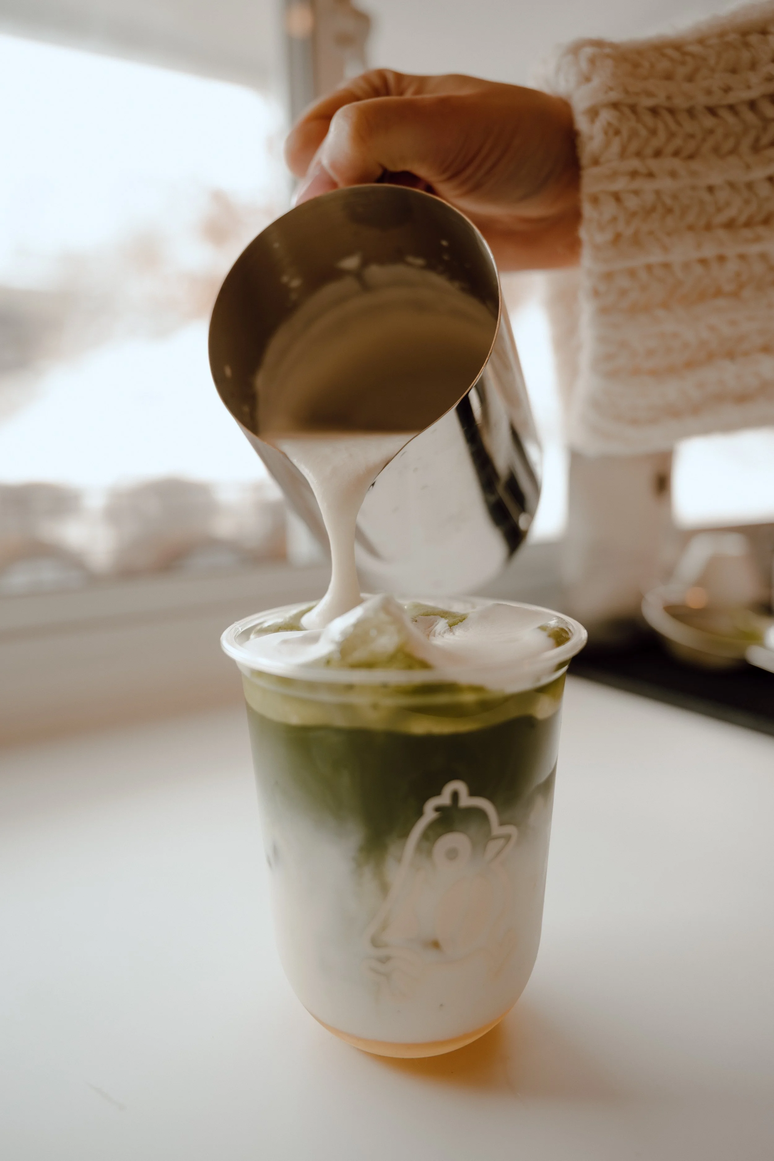 Pouring white cream or foam into a layered matcha latte in a clear cup with a chicken illustration, on a white surface with a window in the background.