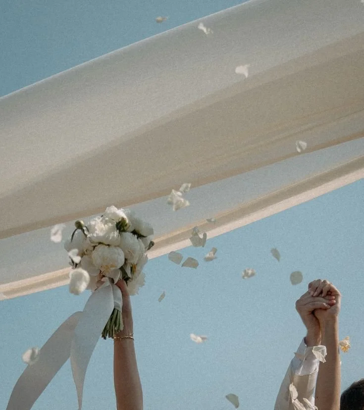 Two people raising their fists, one holding a bouquet of white flowers, under a large white fabric with a clear blue sky in the background.