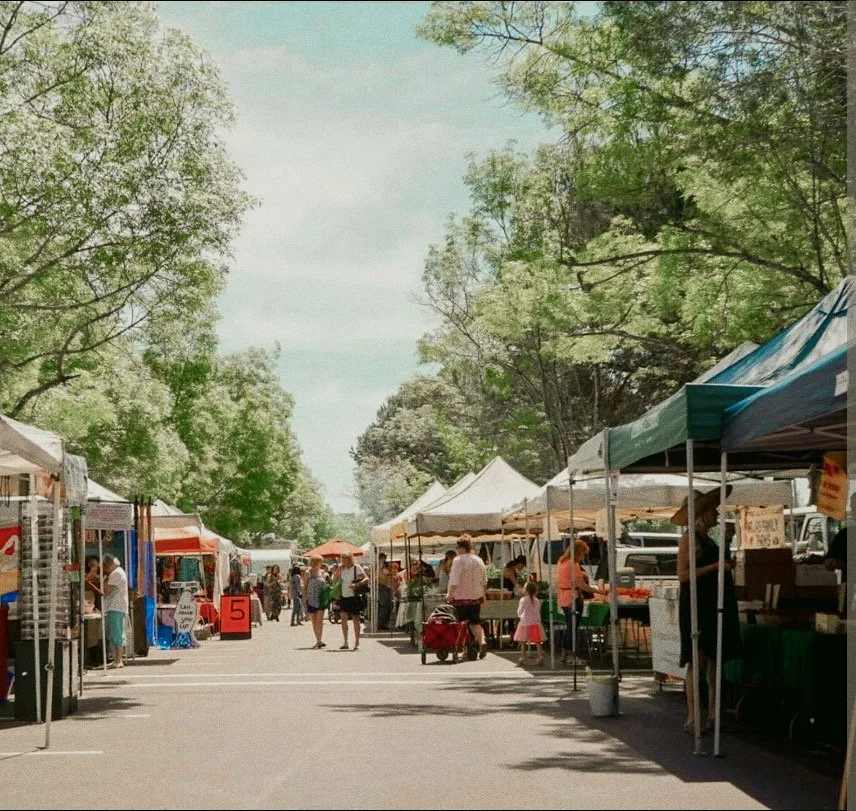 Outdoor market with white tents, people shopping and walking, trees and blue sky in the background.