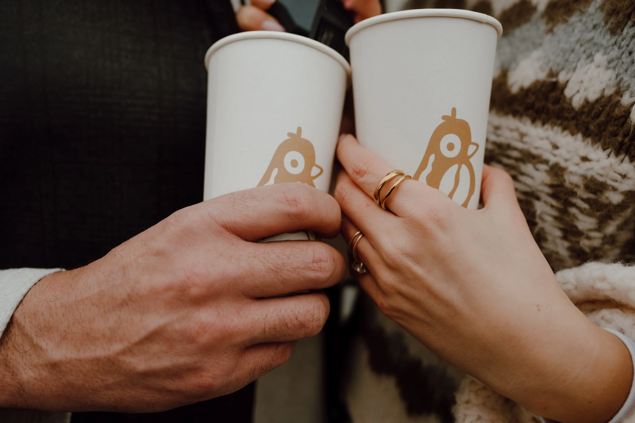 Two people holding white cups with a cartoon bird logo, close-up of hands showing rings, one with multiple rings, the person on the right is wearing a sweater with a patterned design.