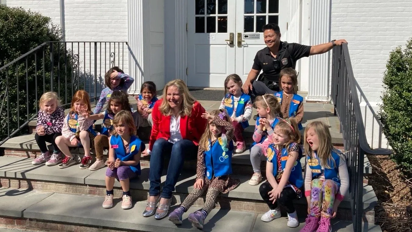 A group of Girl Scouts and two adults sitting on the steps outside a white building. The girls are wearing blue vests, and two adults are sitting with them, one in a red blazer and the other in a black uniform.