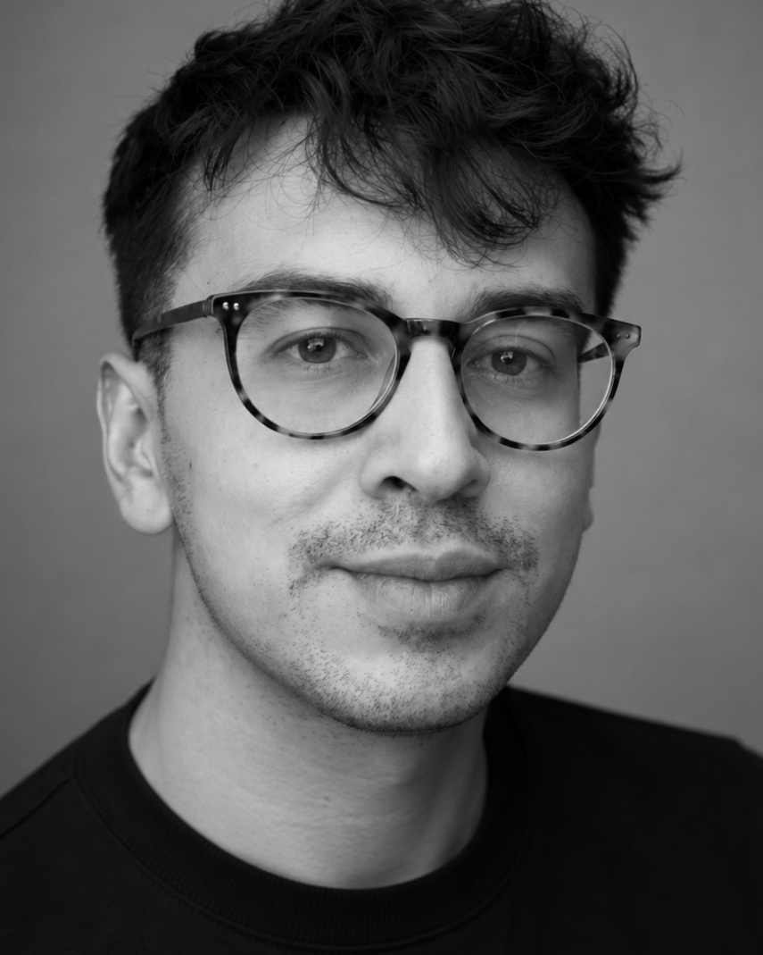 A black-and-white portrait of a young man with dark, wavy hair wearing round, tortoise-shell glasses and a black shirt, looking directly at the camera with a slight smile.
