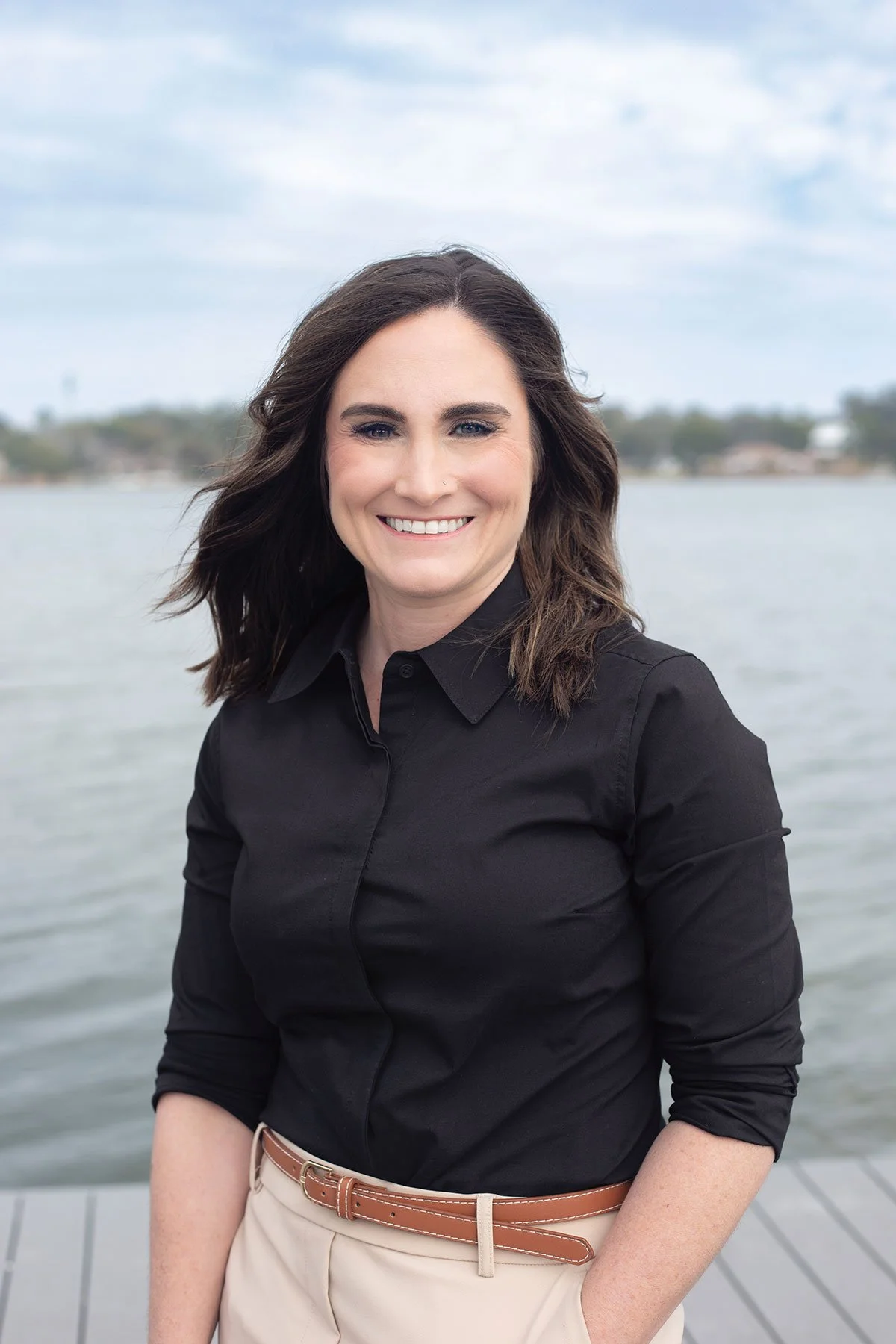 Carissa Smith, LCSW – A woman with shoulder-length dark hair, wearing a black button-up shirt and beige pants with a brown belt, smiling outdoors near a body of water with a cloudy sky in the background.