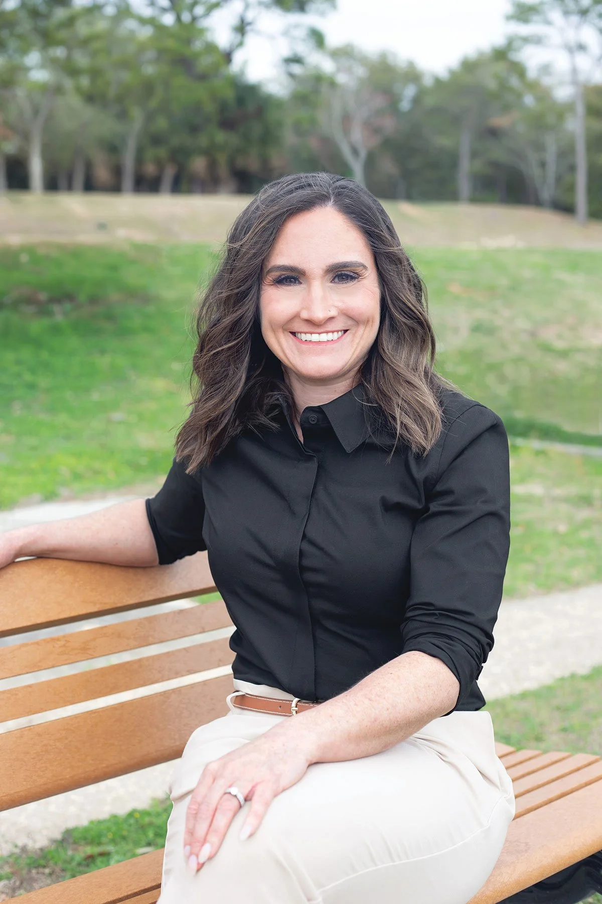 Carissa Smith, LCSW – A woman with brown hair, smiling, wearing a black shirt and light-colored pants, sitting on a park bench with green grass and trees in the background.