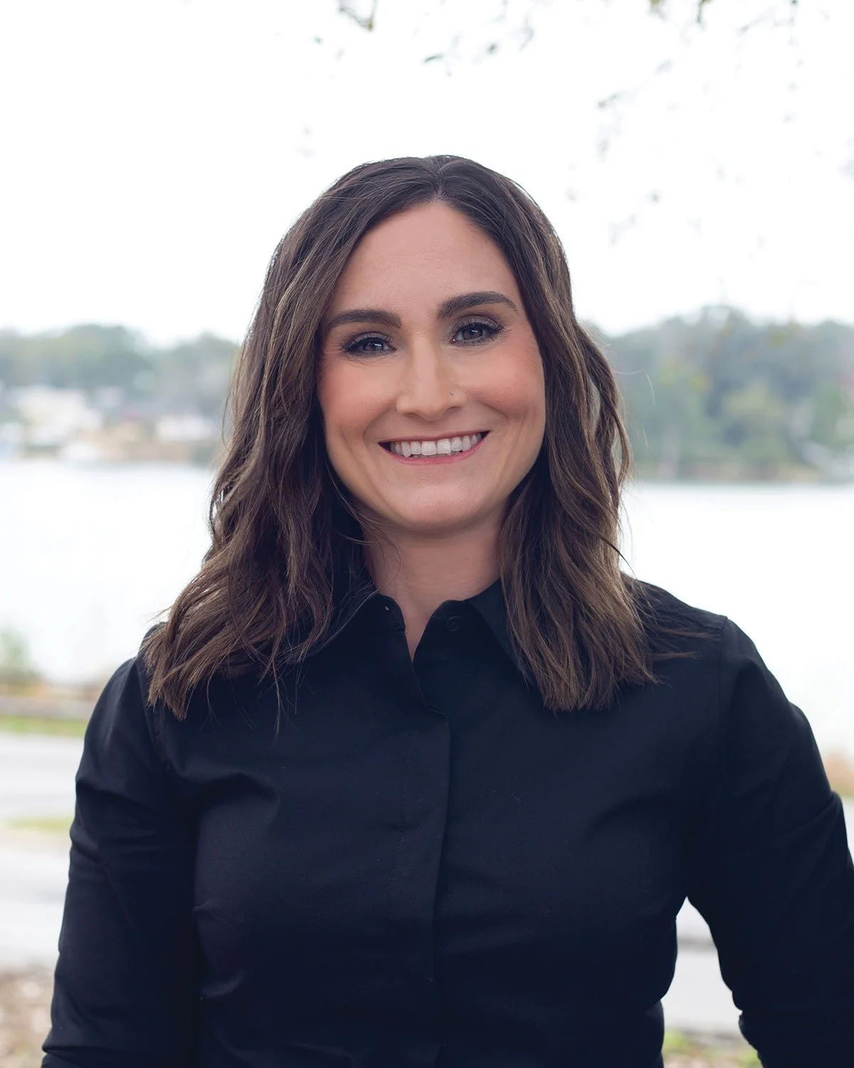 Carissa Smith, LCSW – A woman with shoulder-length wavy brown hair and fair skin, smiling, wearing a black shirt, with a body of water and trees in the background.