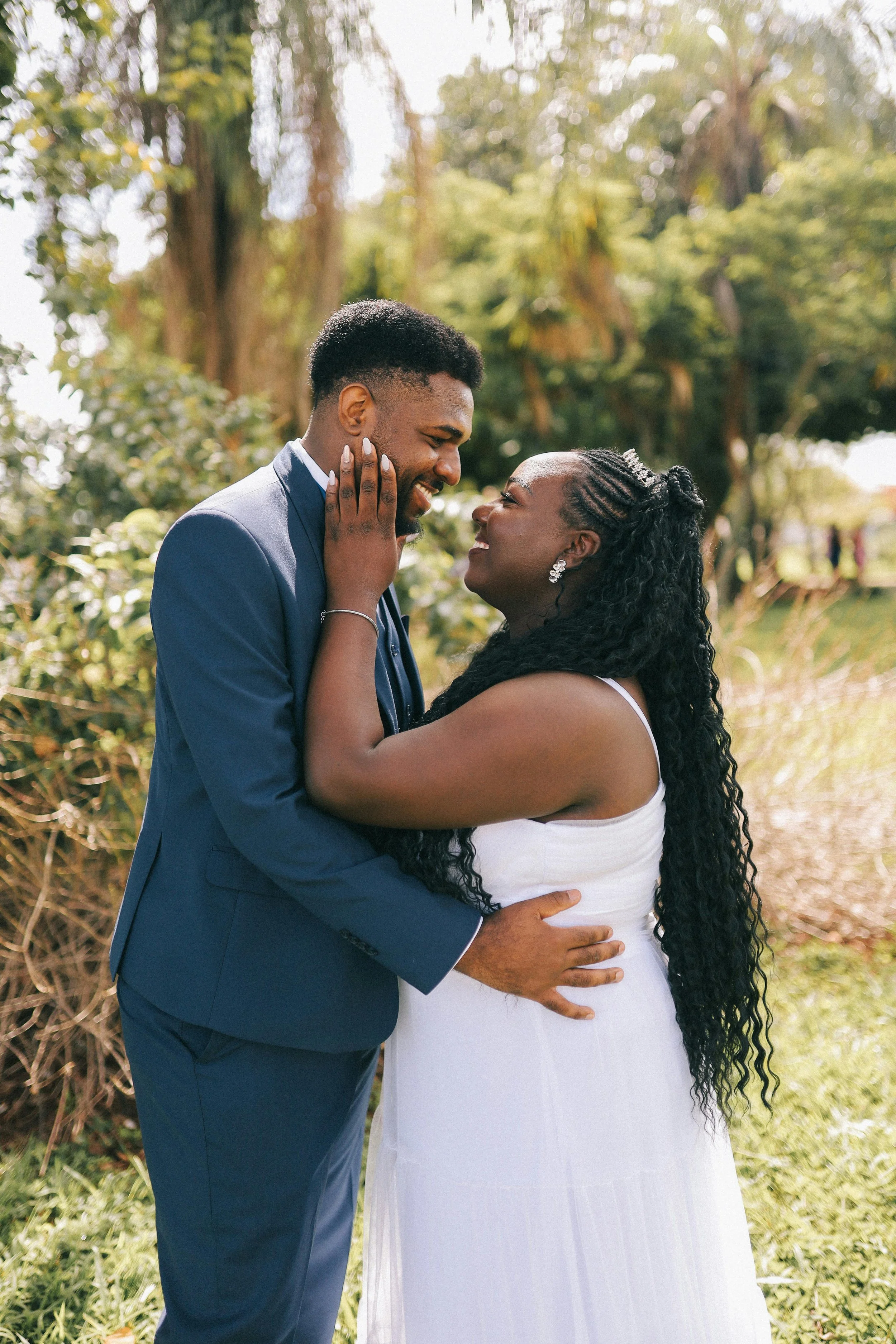 A couple dressed in formal attire embracing outdoors in a lush, green setting.