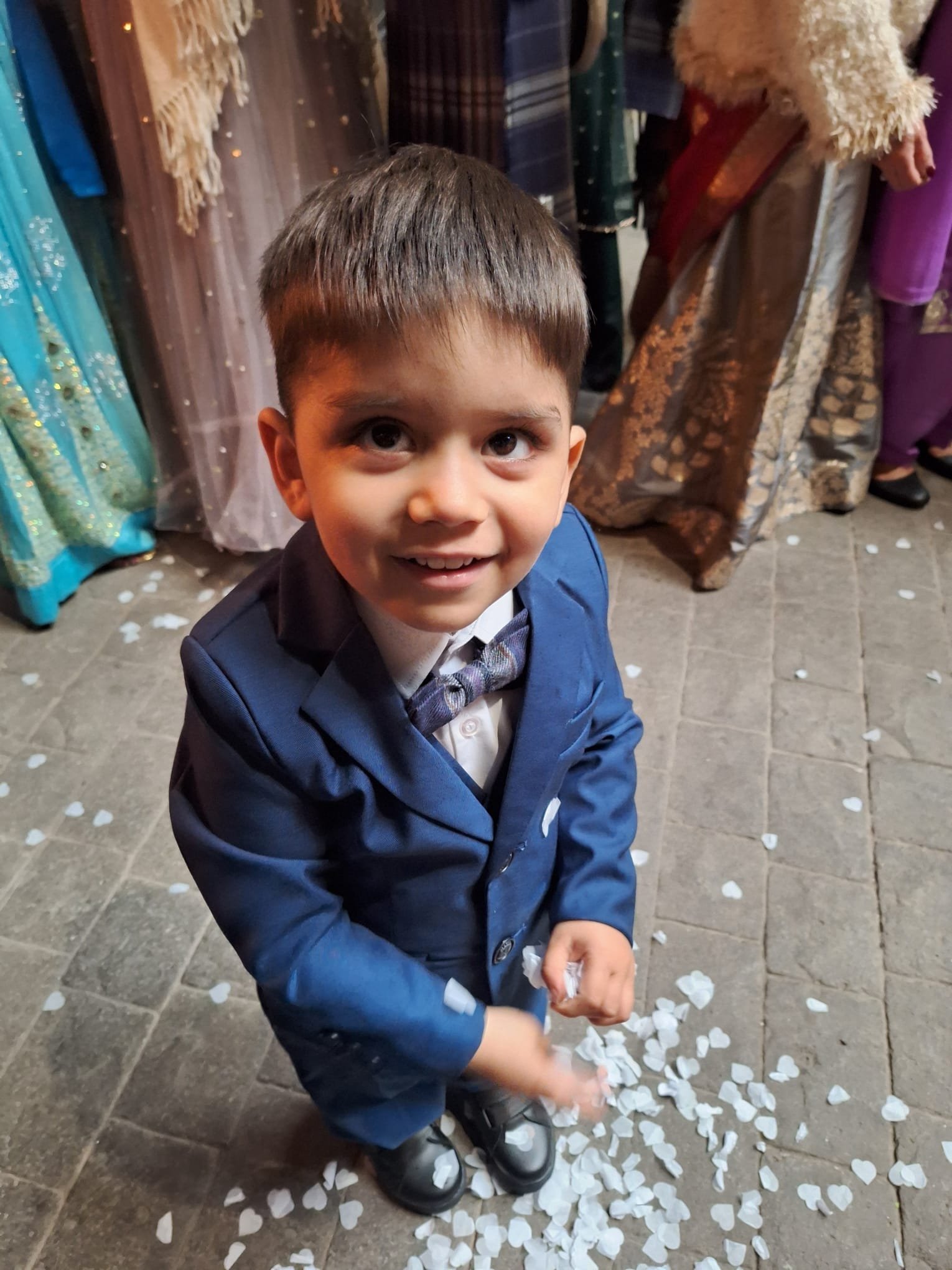 A young boy in a blue suit and bow tie is smiling and looking at the camera while standing on a tiled floor scattered with white flower petals; in the background, people are dressed in formal attire, at an Indian wedding.