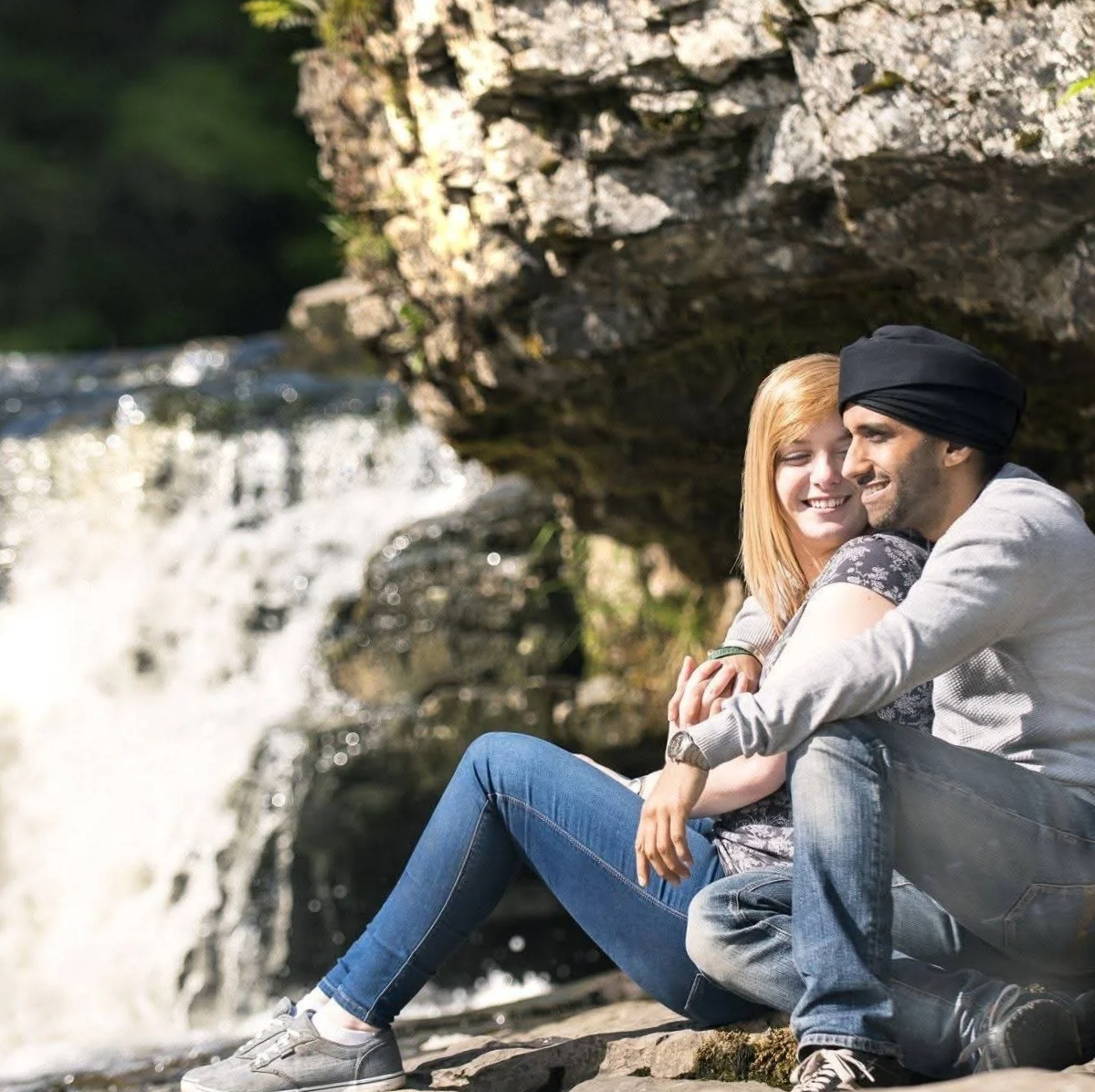 A couple sitting by a waterfall in a natural outdoor setting. The woman has red hair and is smiling, while the man wears a black turban and is also smiling. They are embracing each other.