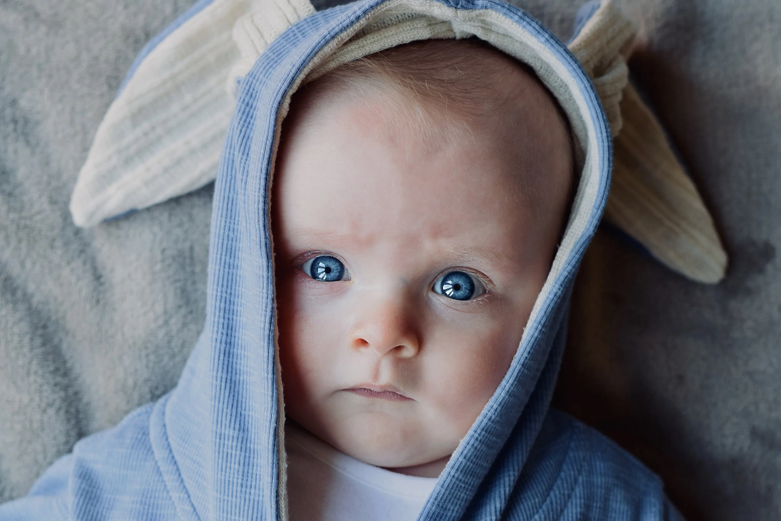 Close-up of a young boy with bright blue eyes, light skin, wearing a hooded jacket with the hood over his head, and a shirt underneath, lying on a gray surface.