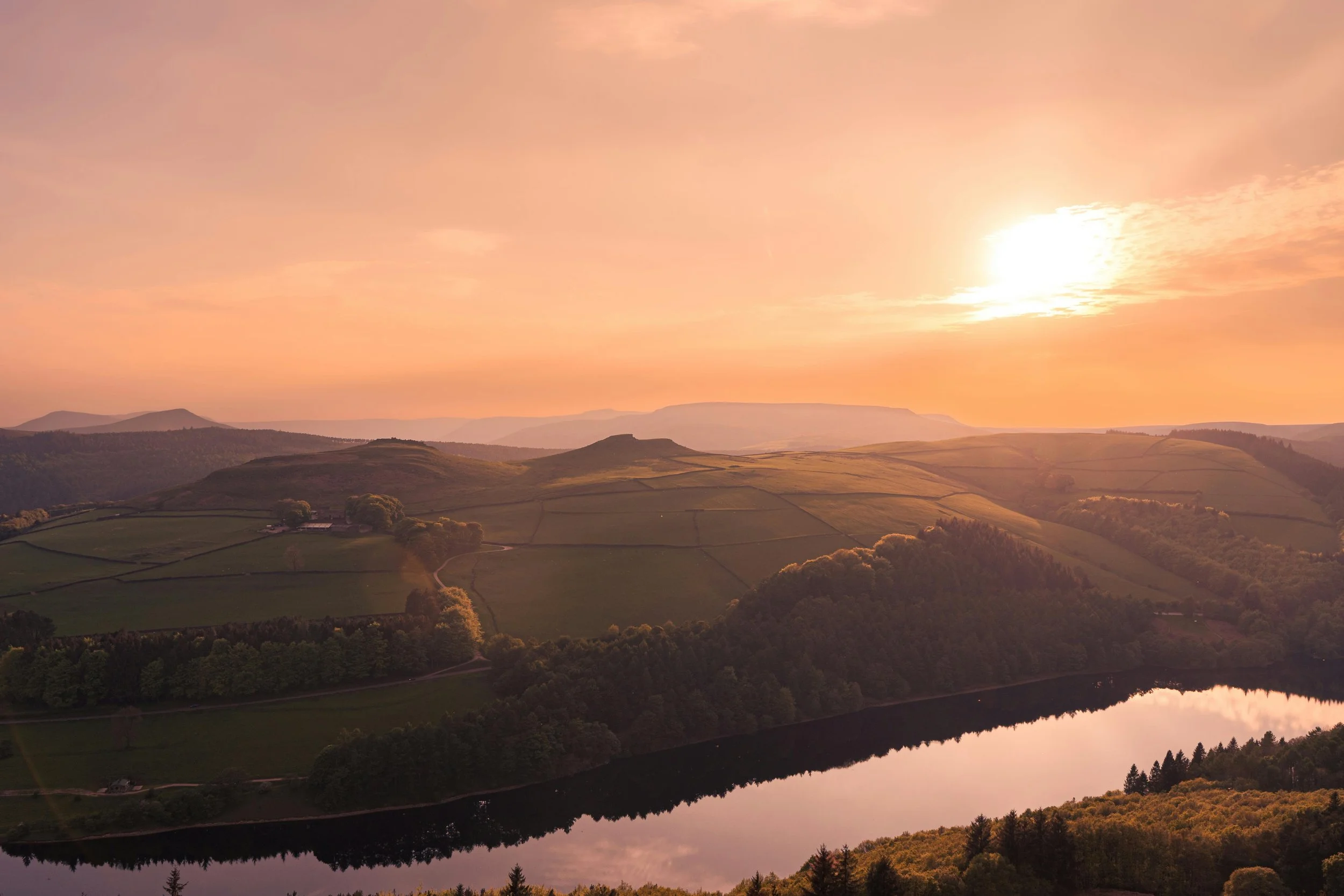 Sunset over rolling hills and a lake reflecting the sky with pink and orange hues.