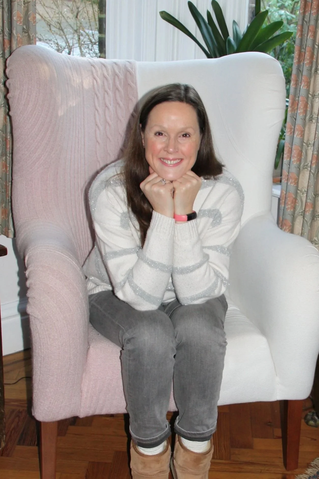 A woman sitting in a white and pink armchair in a room with large windows. There are curtains and plants behind her. She is smiling with her chin resting on her hands.