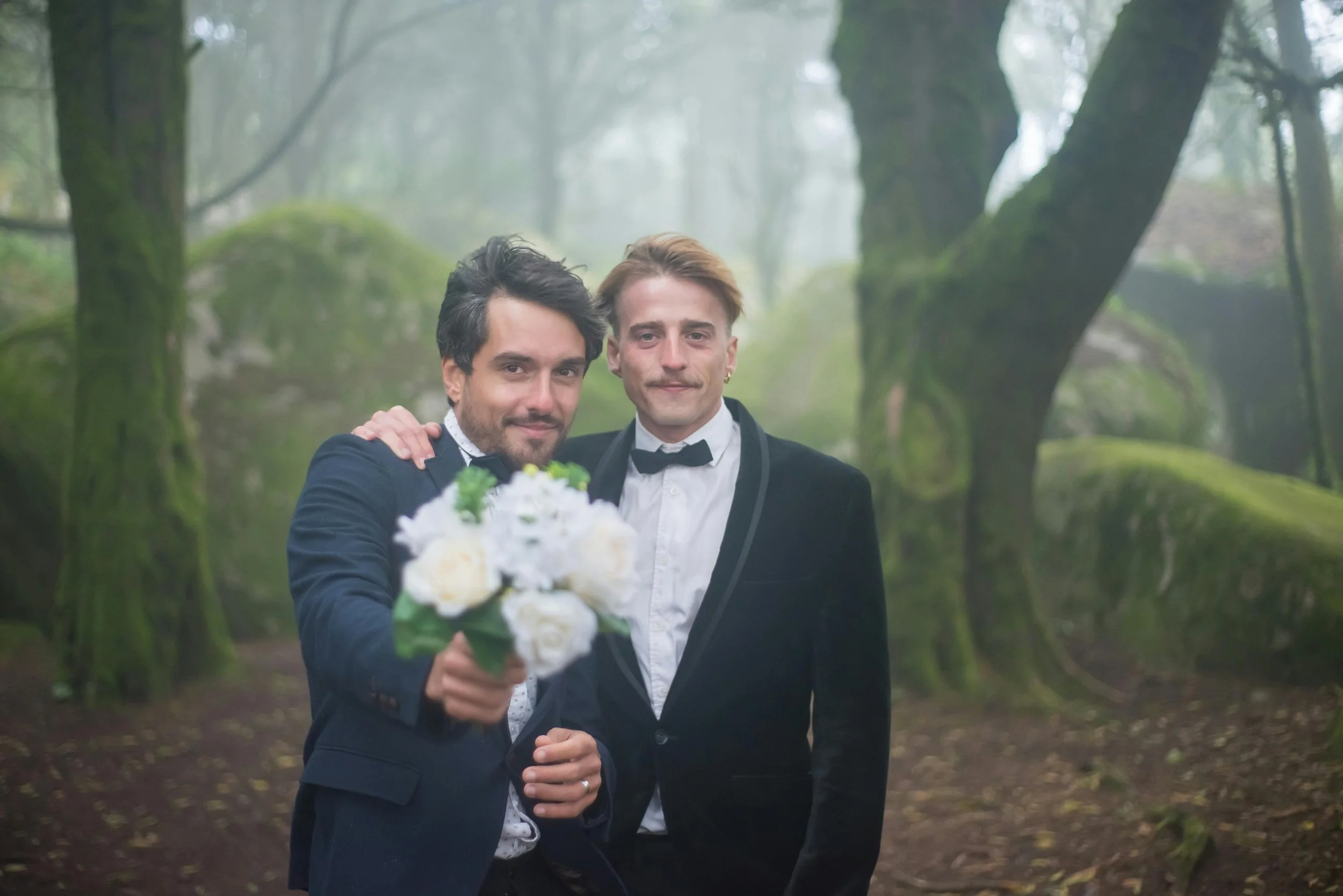 Two men in tuxedos standing in a forest, one holding a bouquet of white flowers, the other with his hand on his husband's shoulder.