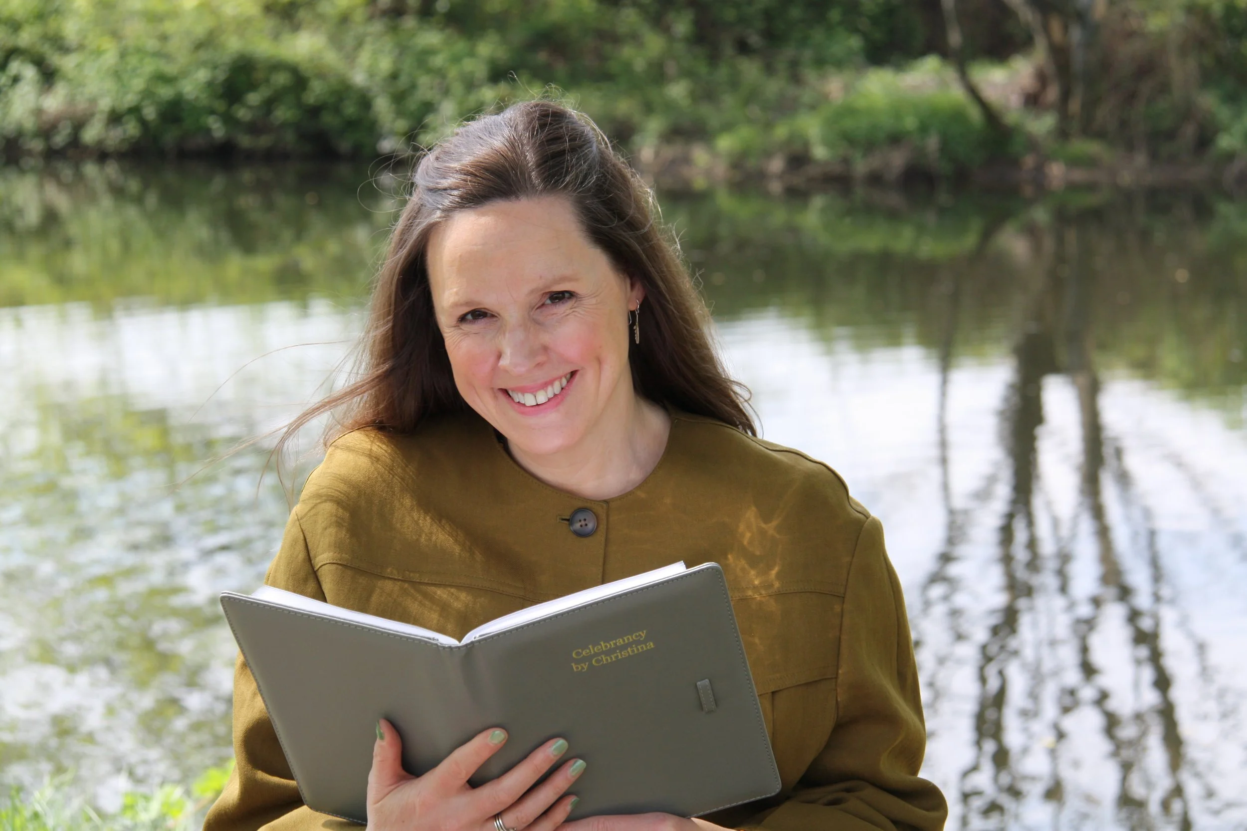 A woman with long brown hair smiling while holding a grey folder and reading outdoors next to a river with green trees in the background, which are reflecting in the river.